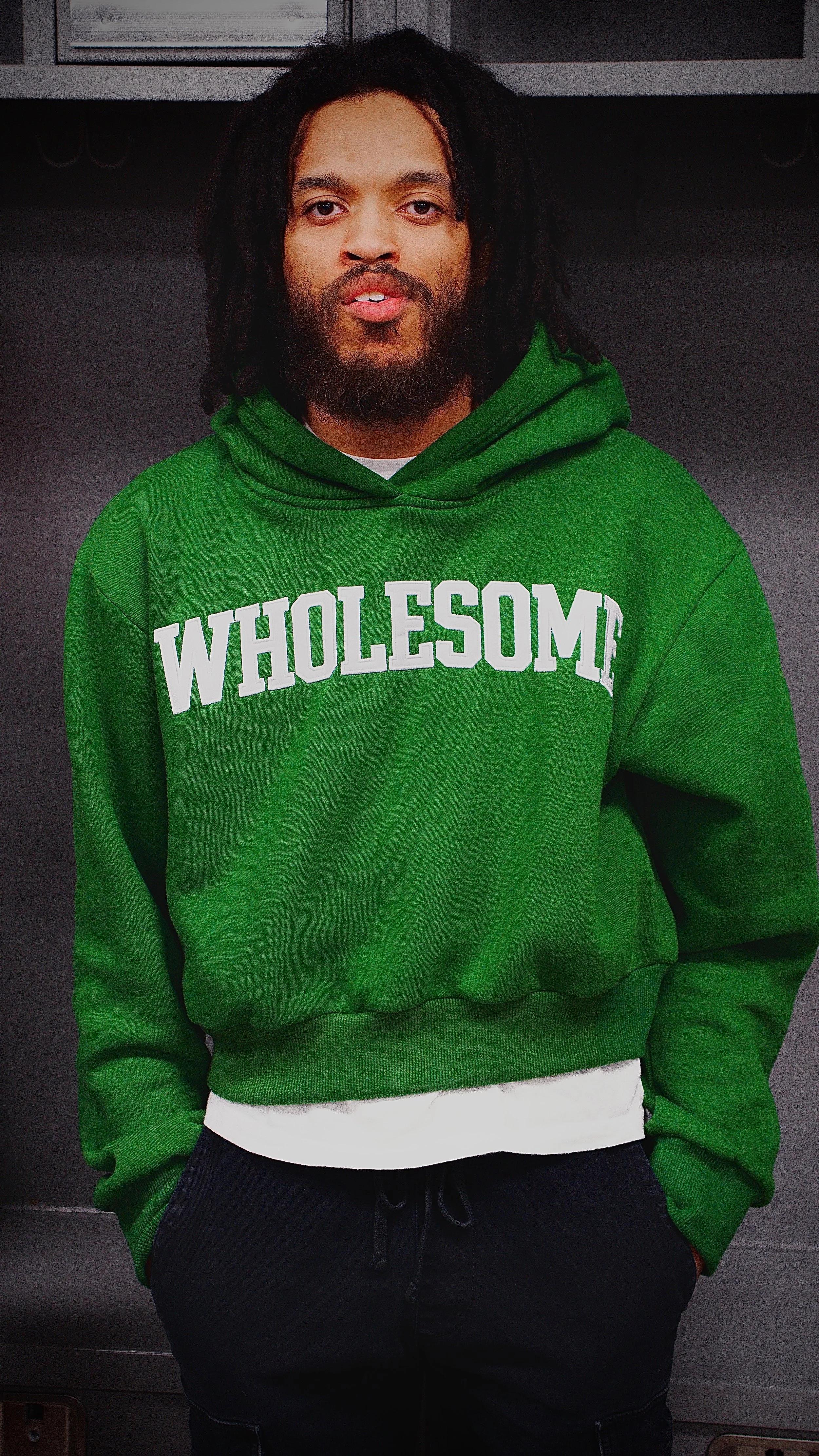 Man with dreadlocks wearing a green hoodie with the word 'WHOLESOME' printed on it and black pants, standing in front of a dark background.