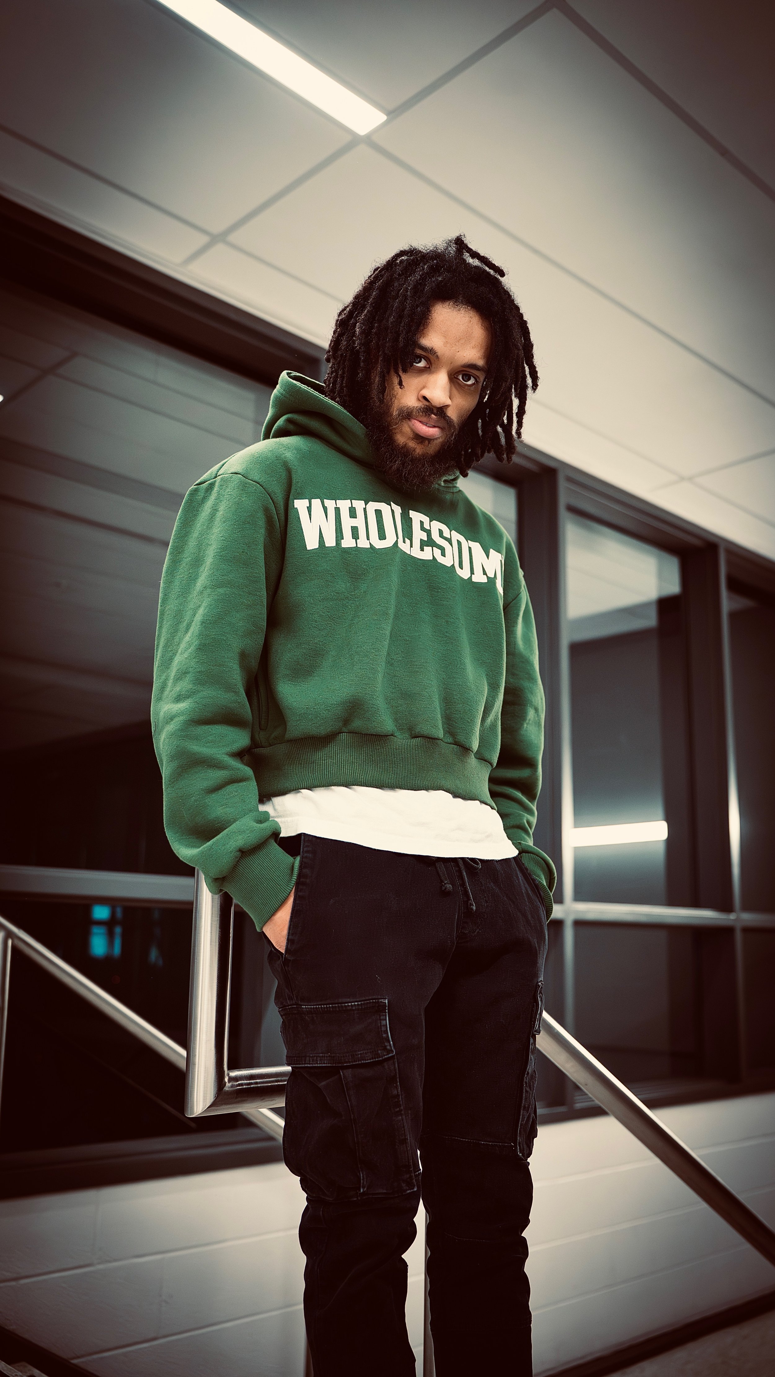 A young man with dreadlocks and a beard, wearing a green hoodie with the word 'WHOLESOME' on it, standing indoors with a modern background of glass windows and metal railings, looking directly at the camera.