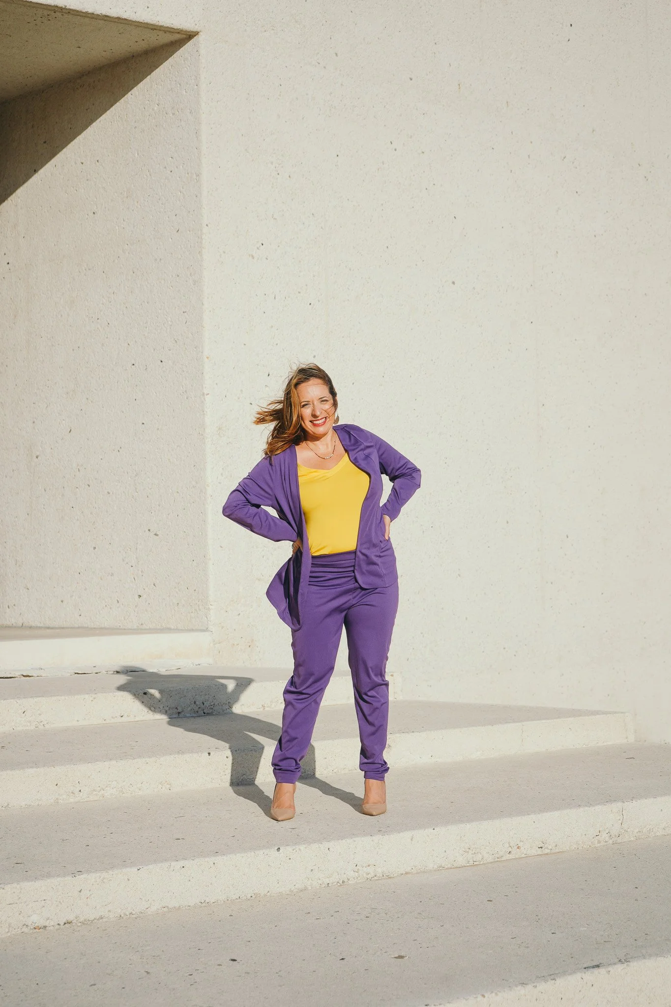 A woman in a purple suit with a yellow top standing outdoors against a plain concrete wall, smiling with her hands on her hips.