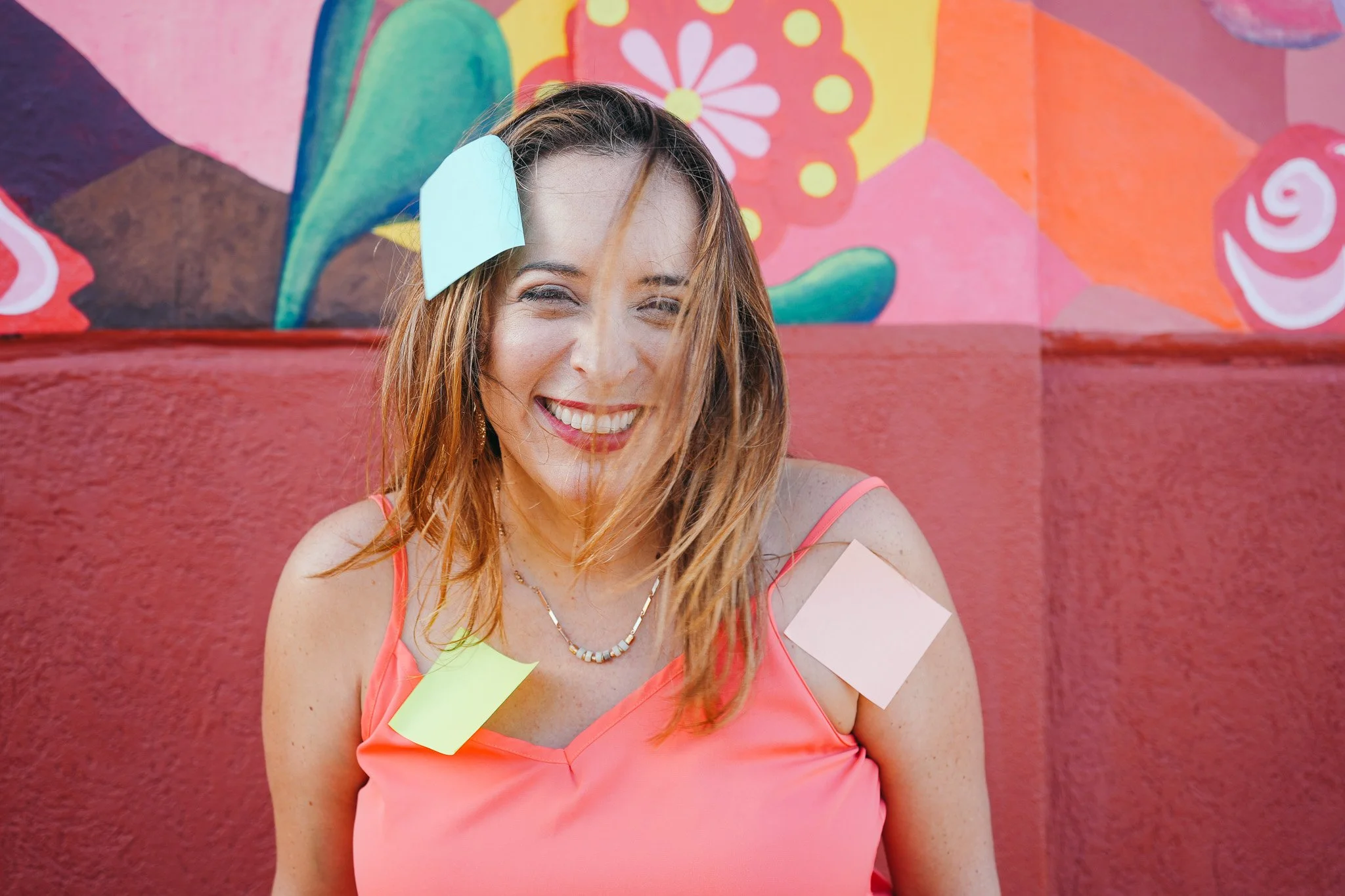 A woman with shoulder-length reddish-brown hair smiling, wearing a coral-colored top and colorful sticky notes attached to her shoulder, chest, and forehead, in front of a bright, colorful mural.