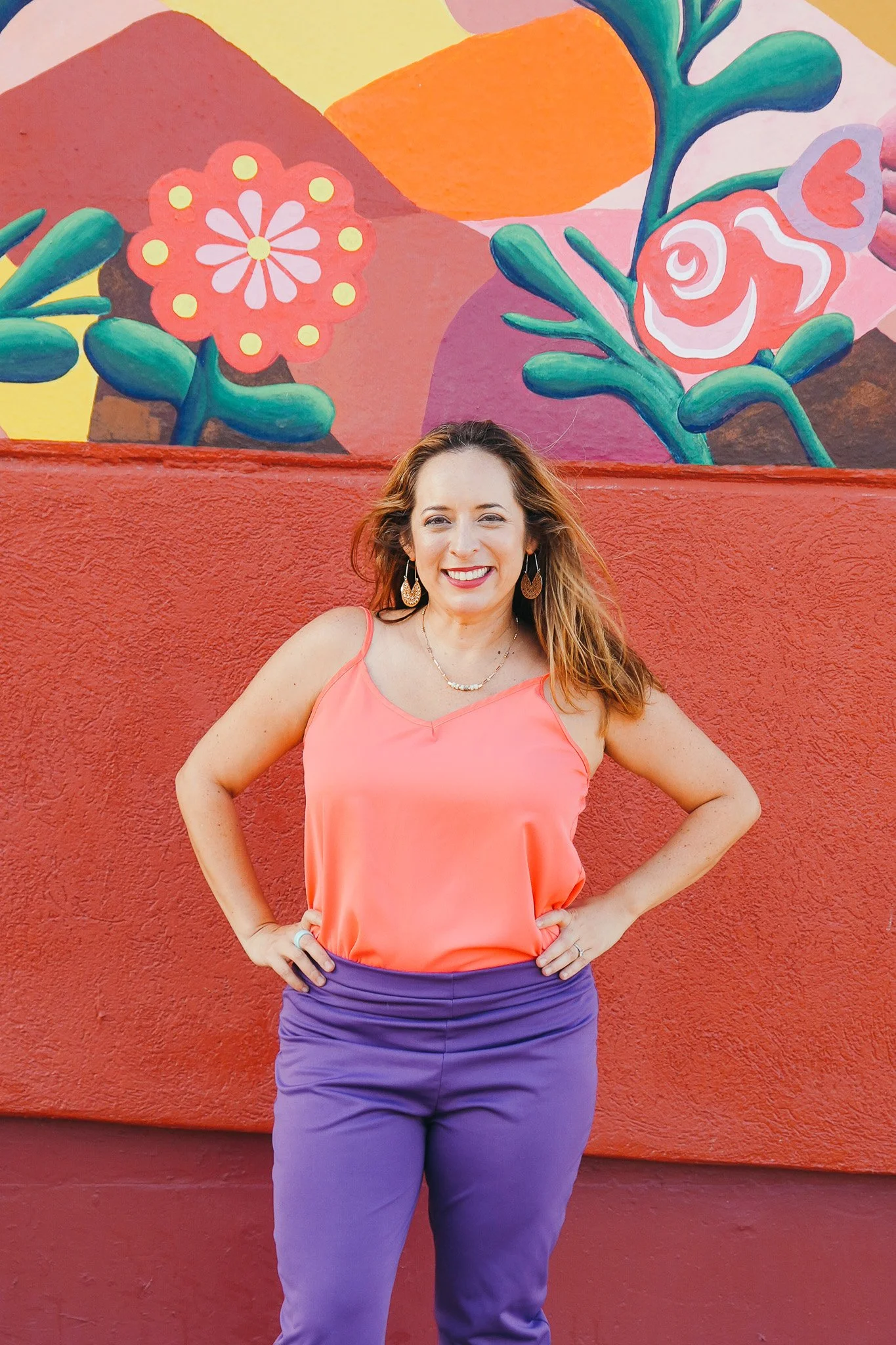 A woman with light skin and long, wavy hair wearing a coral sleeveless top, purple pants, and jewelry, smiling in front of a colorful mural with abstract floral designs.