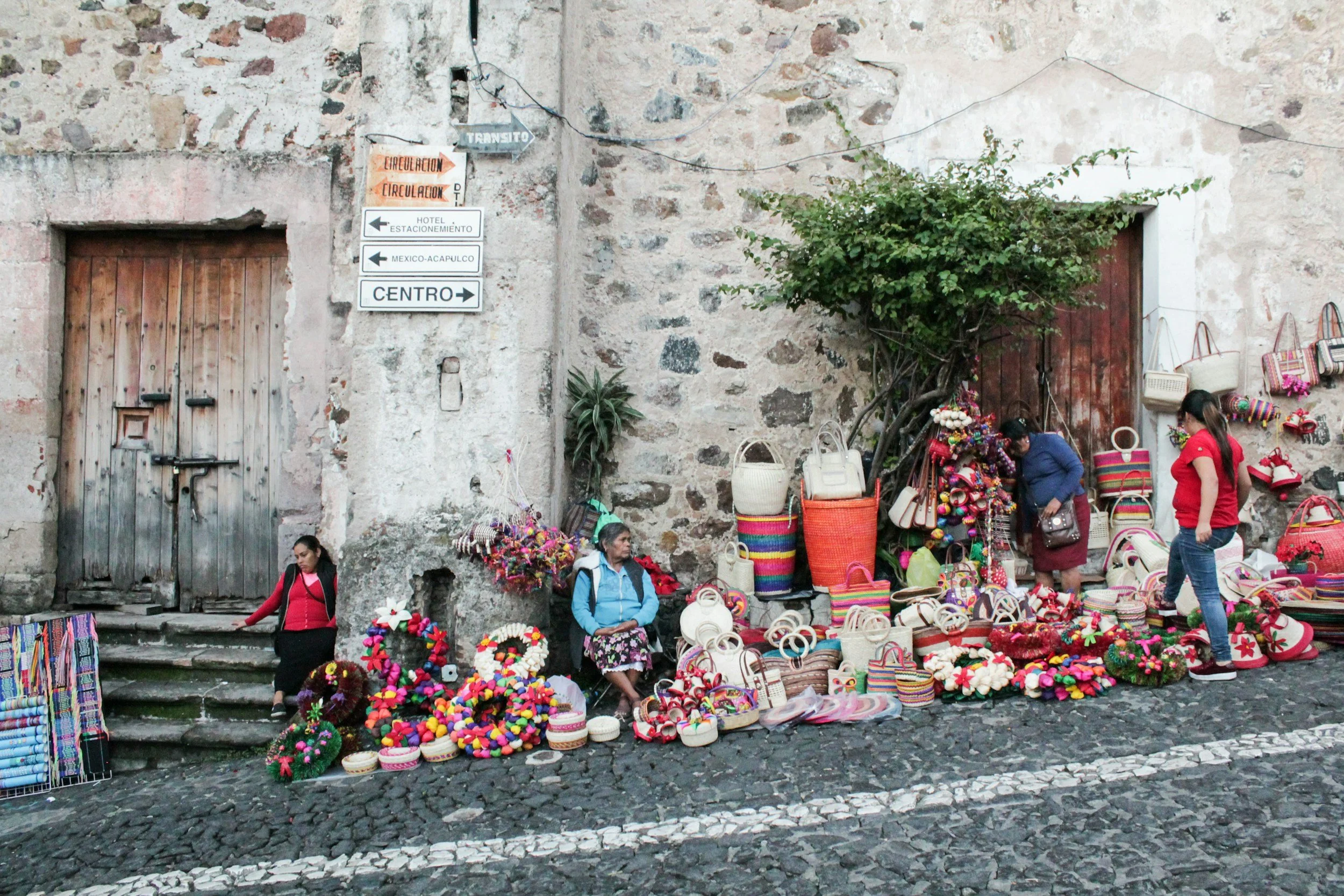 Street vendor selling colorful woven bags, hats, and wreaths outdoors against a weathered stone wall, with three women browsing and selling, and signs pointing to various city directions.