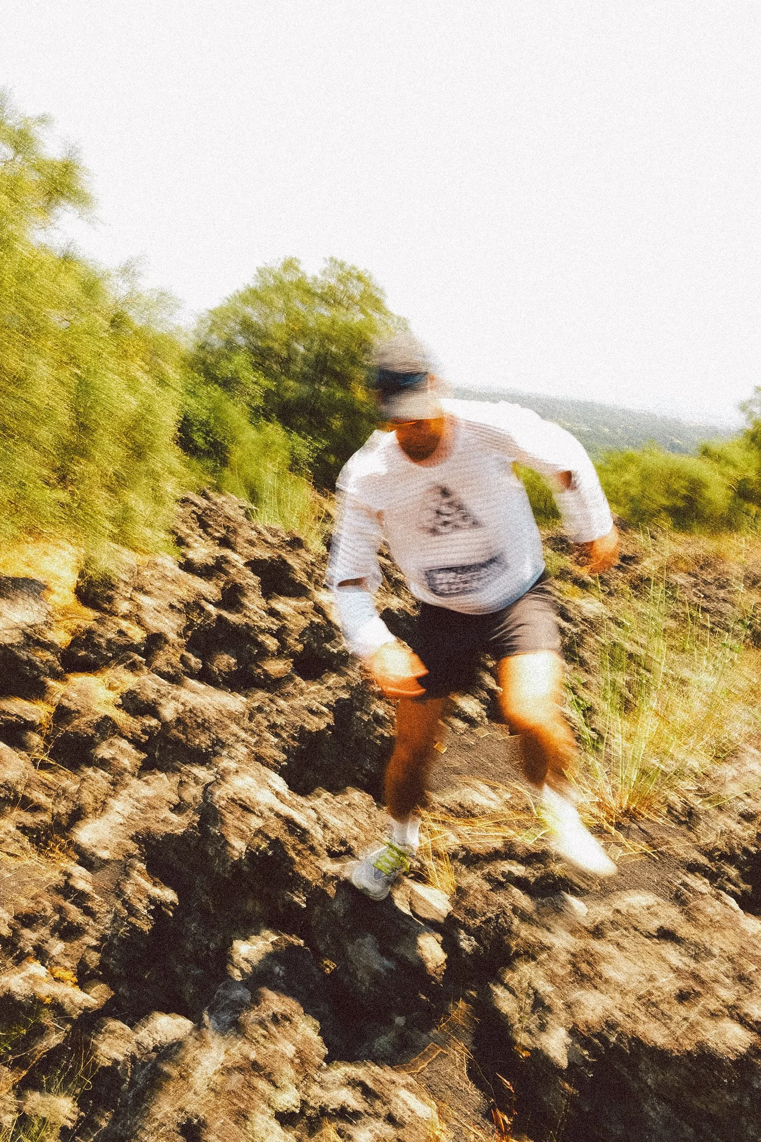 A person wearing a white long-sleeve shirt, shorts, and a hat, walking along a rocky trail with greenery on the sides and a distant view of hills or mountains.