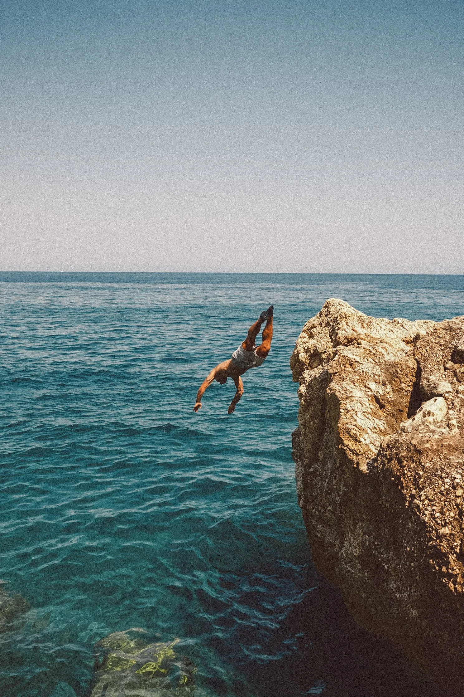 A person in swim trunks is performing a handstand on top of large rocks by the ocean, with the sea and a clear sky in the background.