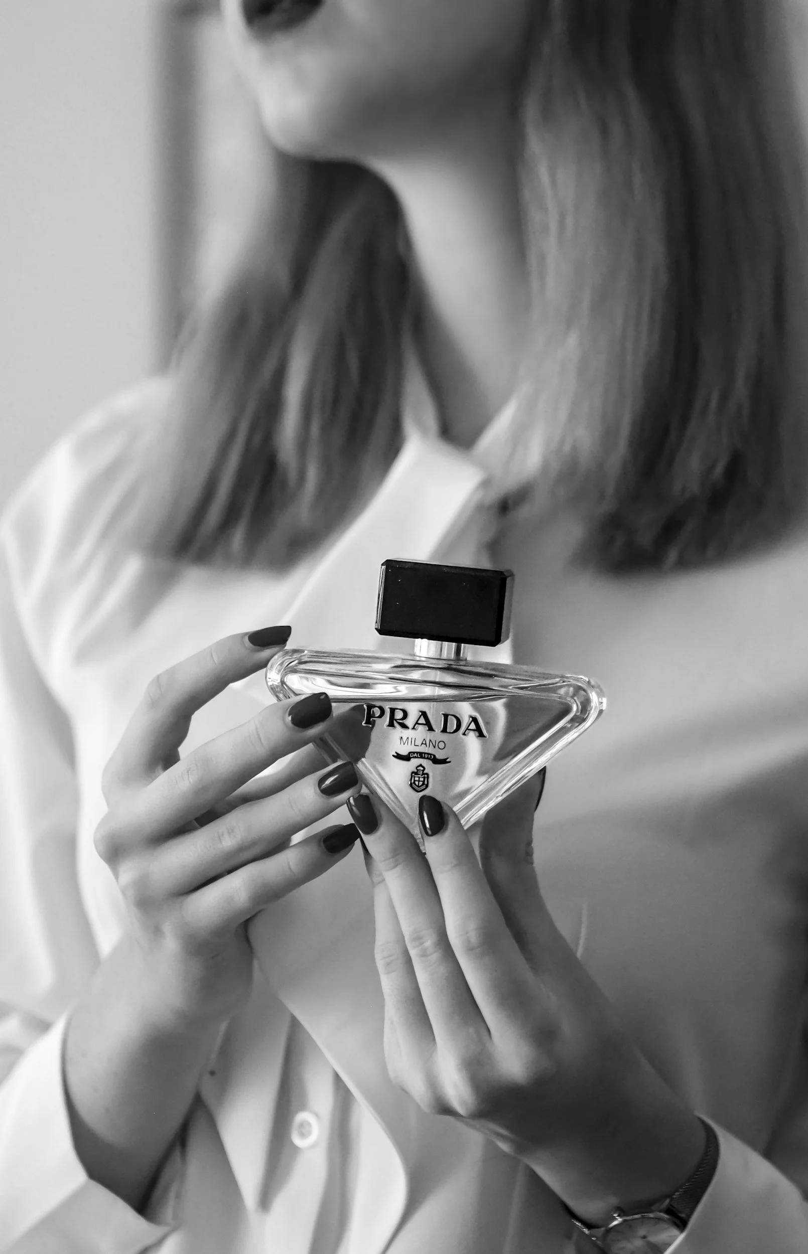 A woman holding a Prada perfume bottle in her left hand, with painted fingernails, wearing a long-sleeve shirt, with single bracelet and watch on her wrist. The photo is in black and white.