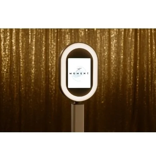Photobooth with illuminated oval frame, gold sequin curtain background, digital screen displaying the word 'MOMENT' and a smiley face icon.