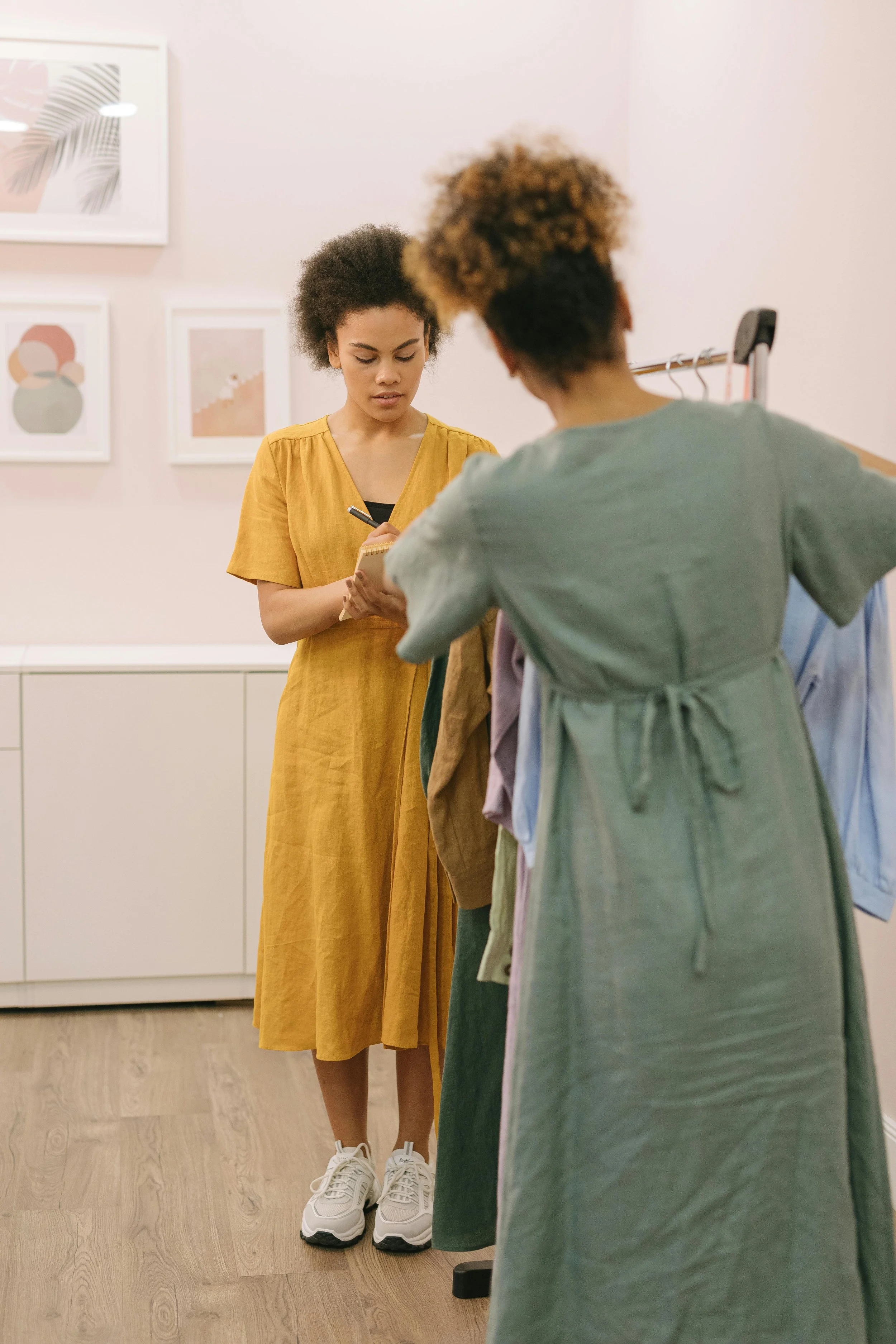 Two women shopping for clothes in a store, one is holding a notepad and pen, the other is browsing clothes on a rack.
