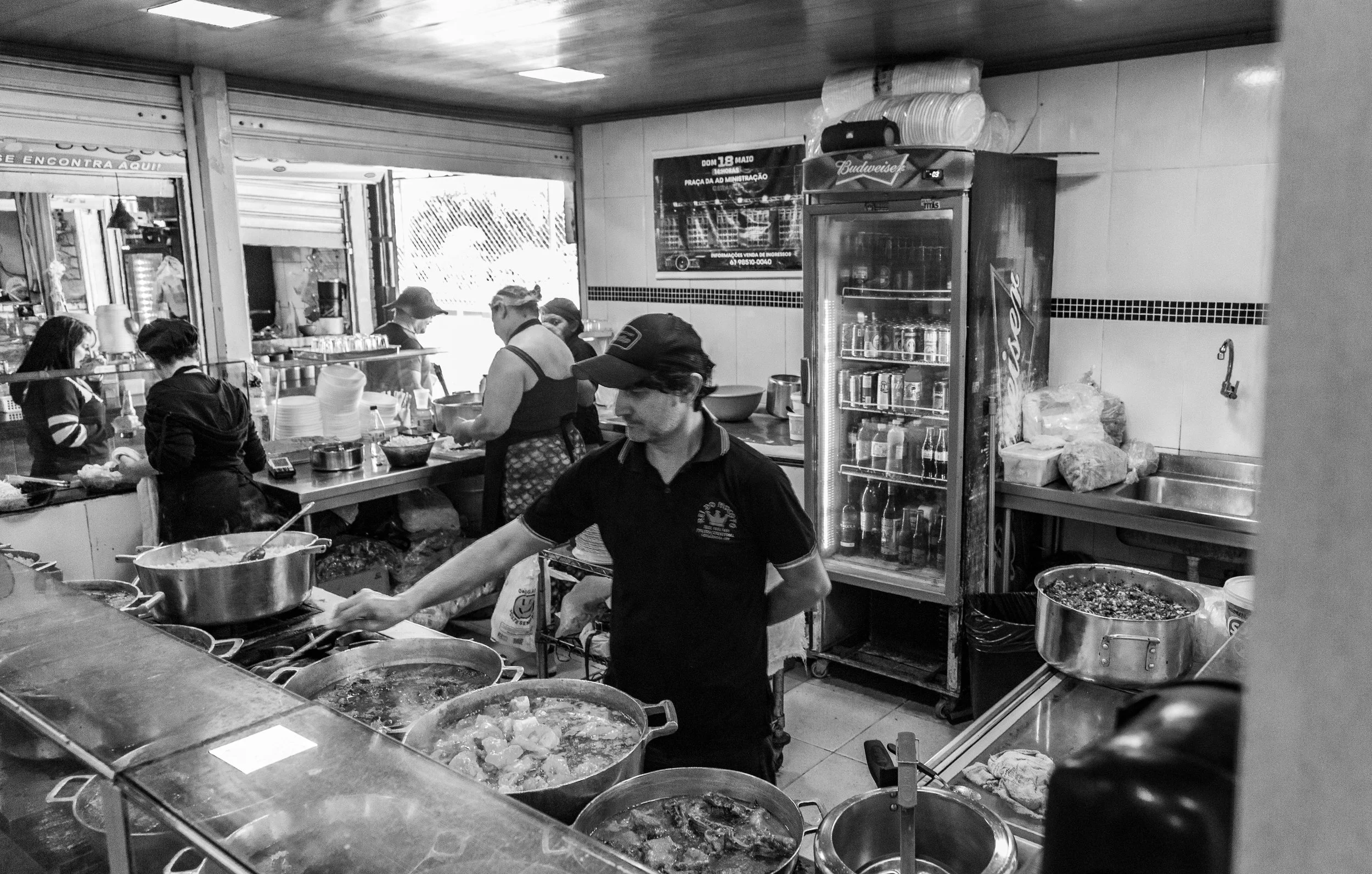 A busy kitchen in a restaurant with several workers cooking and preparing food. There are large pots on the stove, a refrigerator filled with drinks, and various kitchen supplies visible.