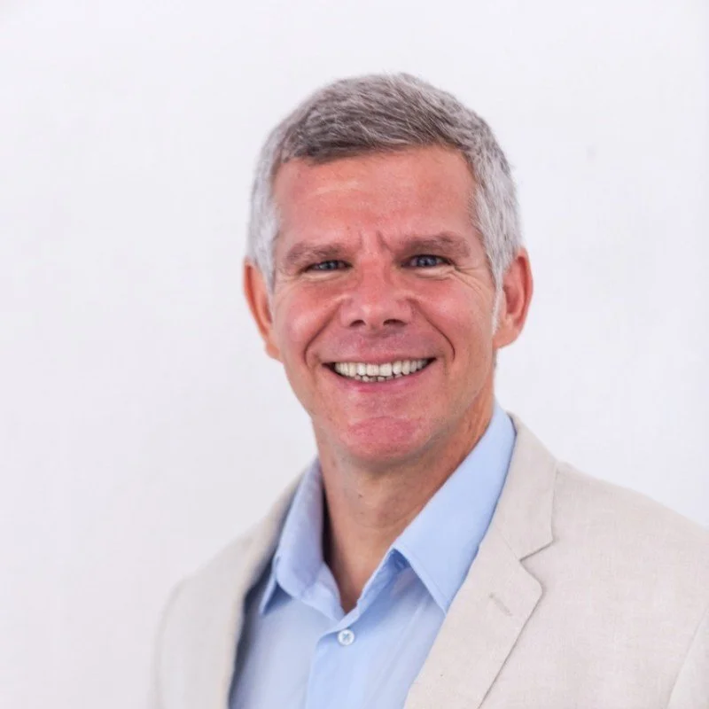 A middle-aged man smiling, wearing a beige blazer over a light blue shirt, with short gray hair, standing against a plain white background.