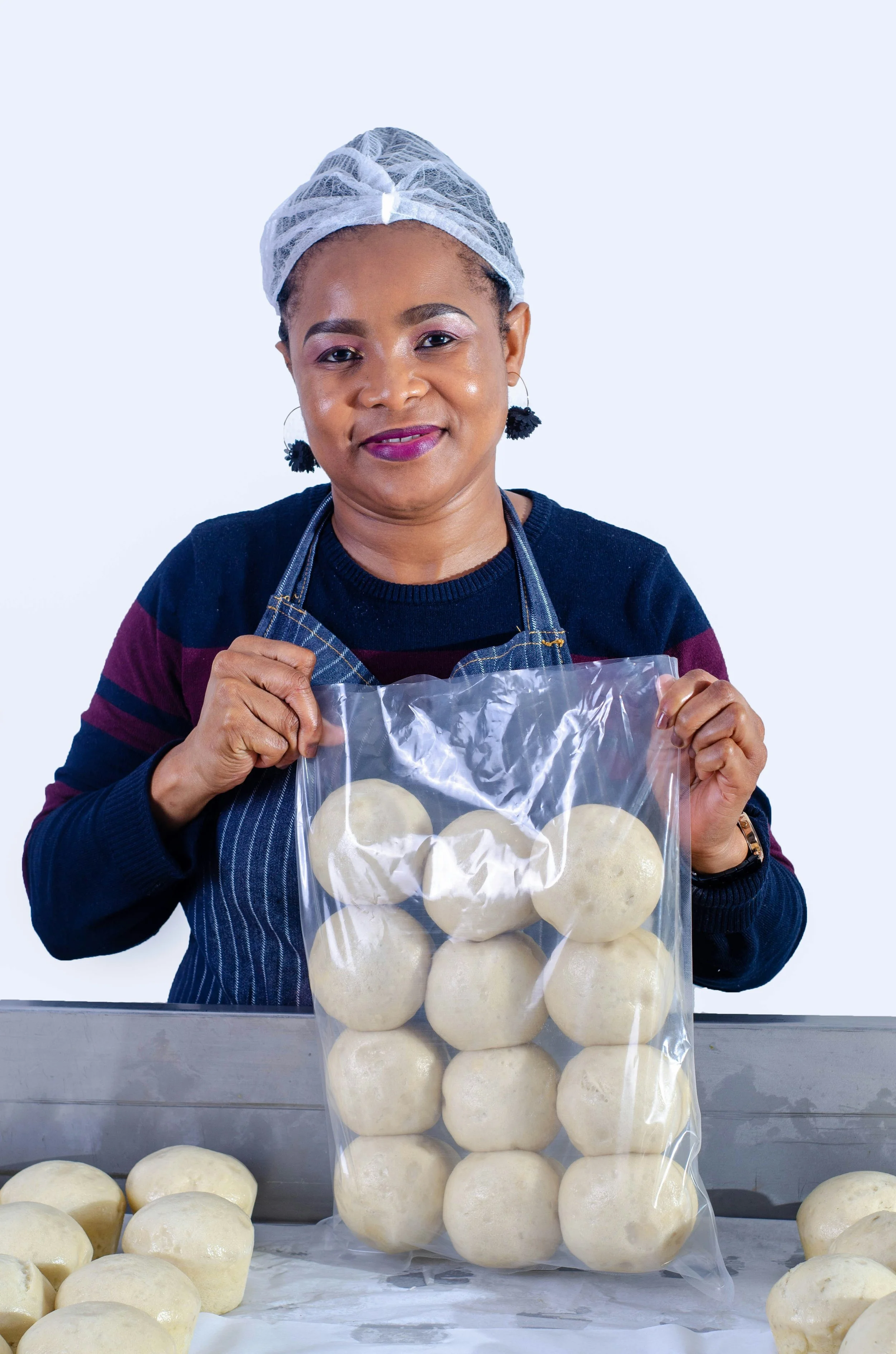 A woman wearing a hairnet, earrings, a striped sweater, and an apron holding a plastic bag filled with round dough balls, standing behind a table with dough balls.