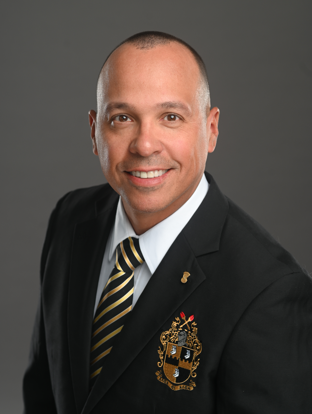 Man in a black suit with a crest on the blazer, striped tie, smiling, plain dark background