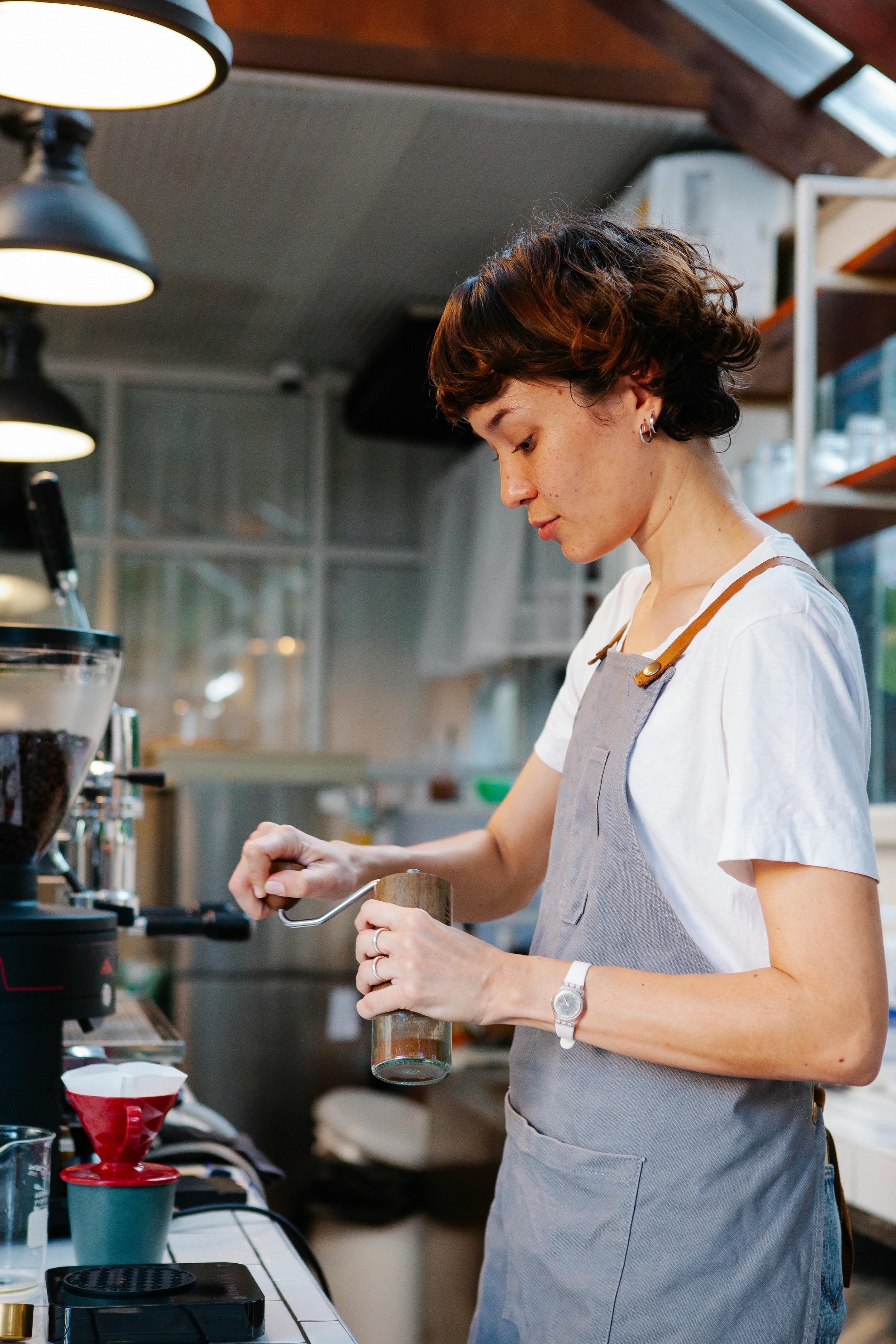 A woman in a kitchen preparing coffee, using a manual grinder, surrounded by coffee equipment and supplies.