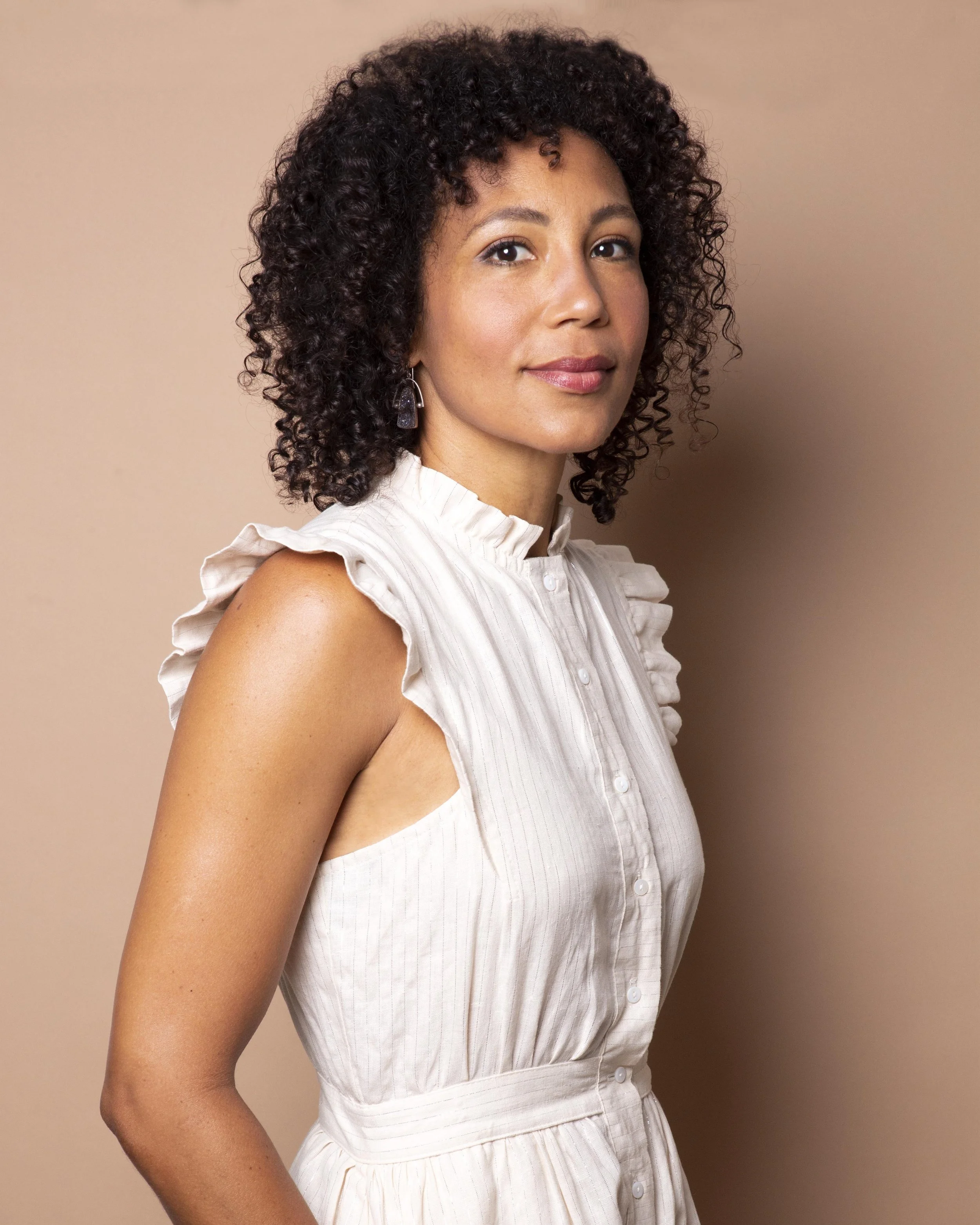A woman with curly dark hair wearing a white sleeveless blouse with ruffled shoulders, standing against a beige background.