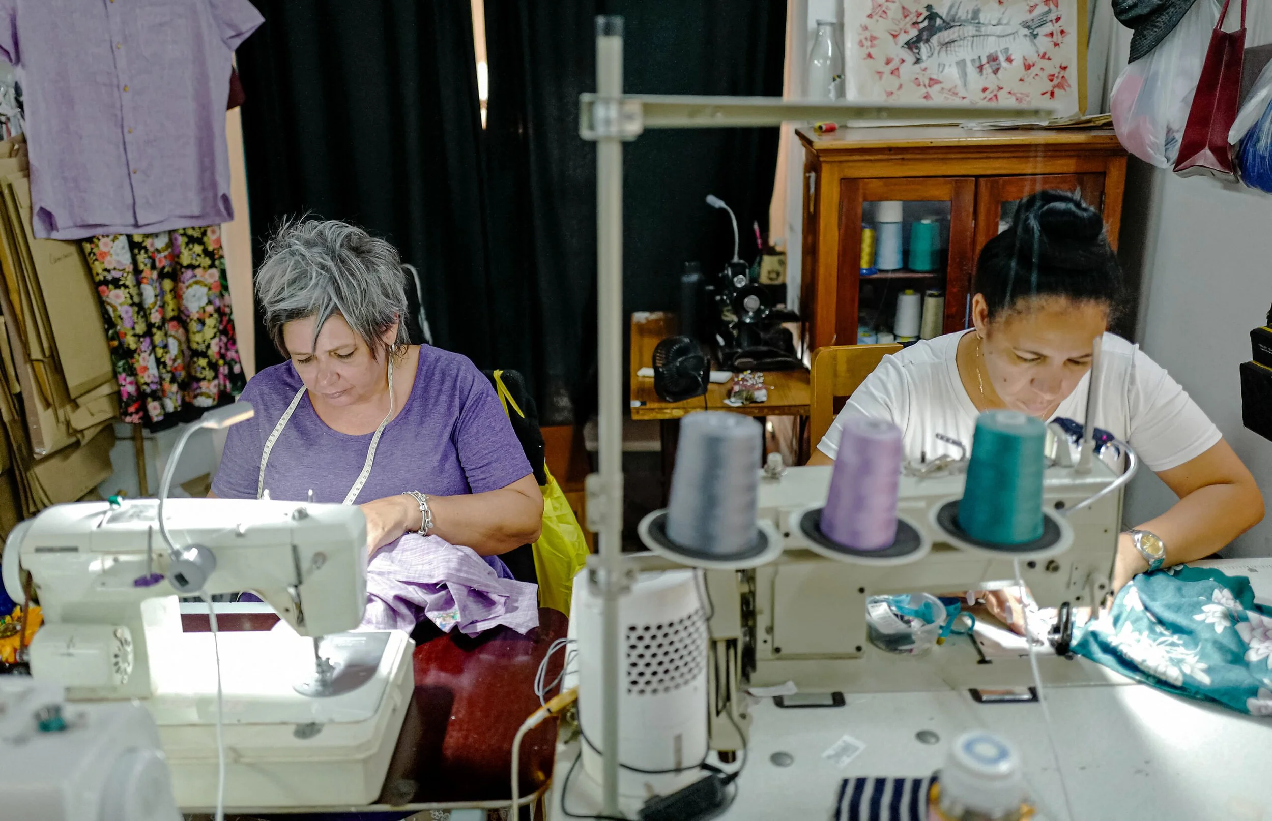 Two women working with sewing machines in a textile workspace, surrounded by colorful threads and fabrics.