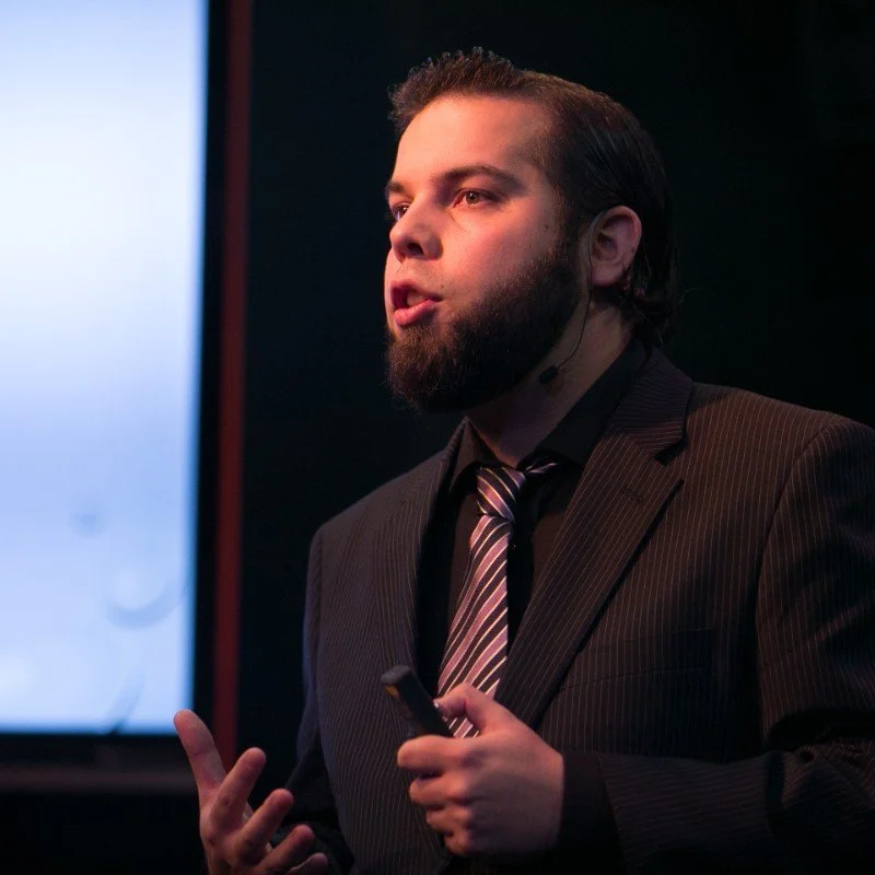 A man with a beard and dark hair wearing a pinstripe suit and striped tie, speaking and gesturing with his hand while holding a remote control in a darkened room with a large screen behind him.