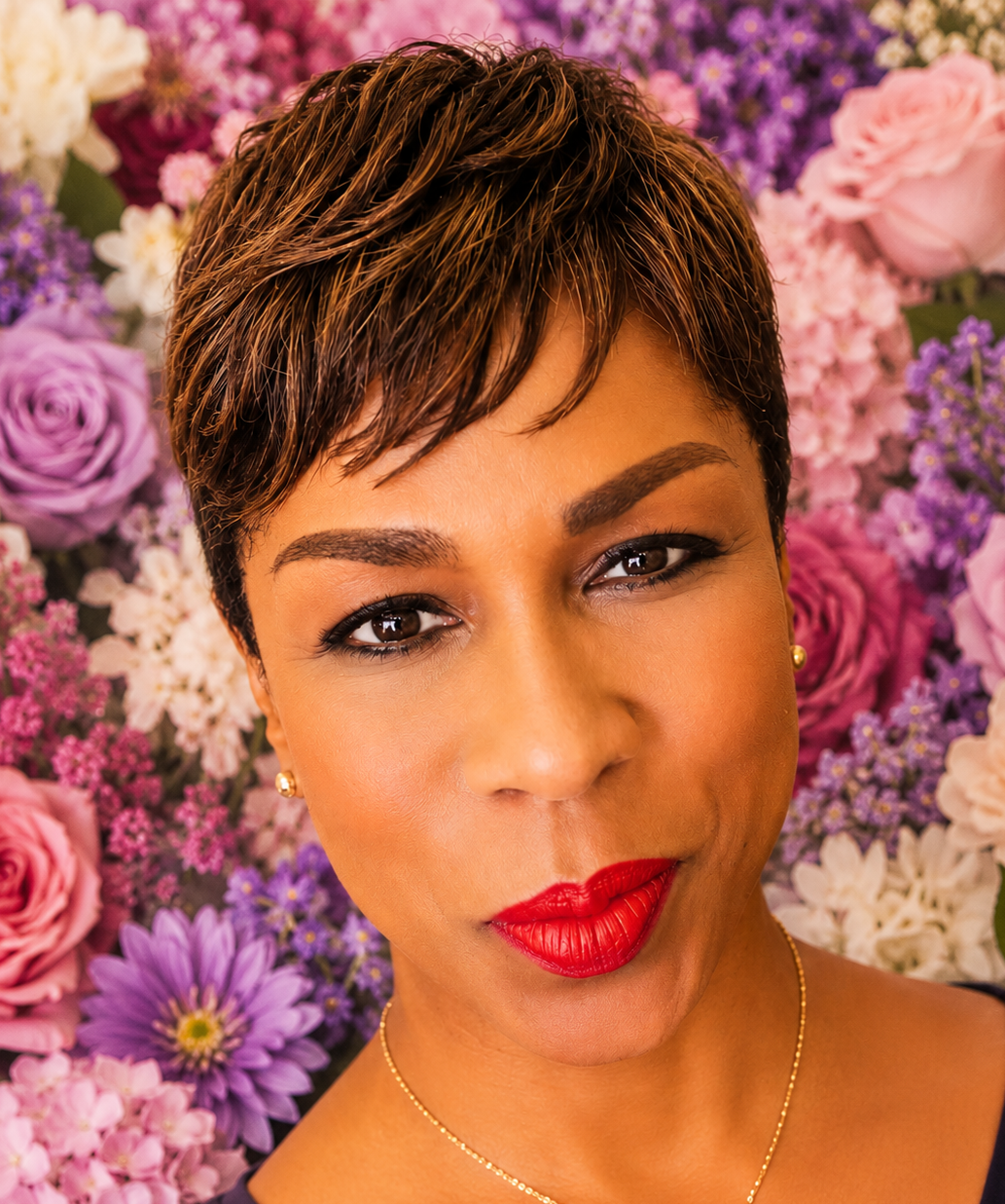 Close-up portrait of a woman with short, textured brown hair, red lipstick, and gold earrings, in front of a colorful background of pink, purple, and white flowers.