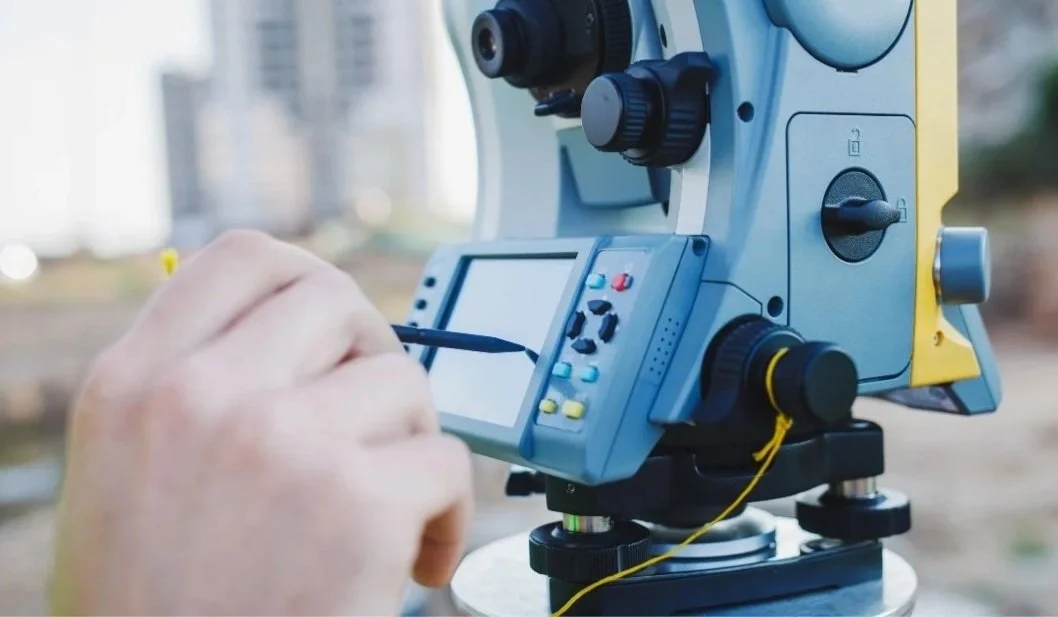 Close-up of a person's hand adjusting a control panel on a theodolite mounted outdoors in an urban area.