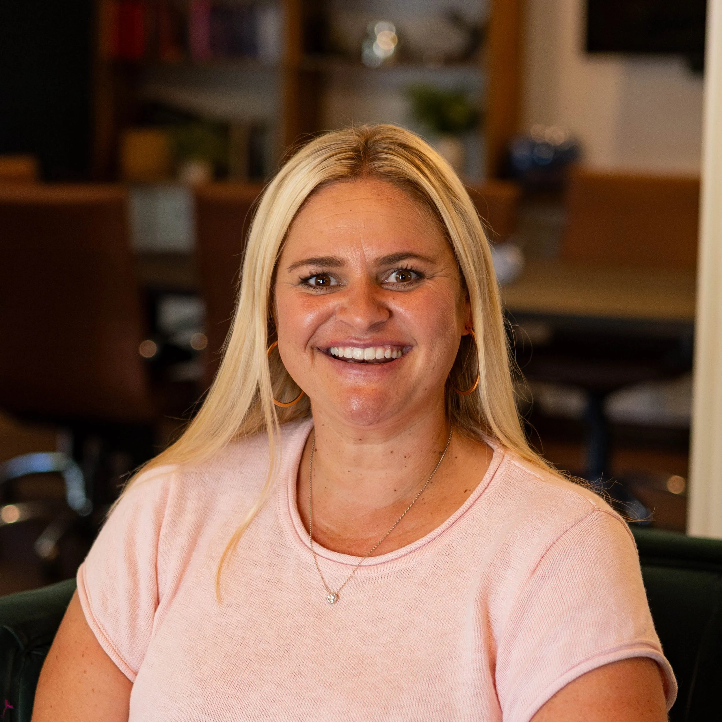 Smiling woman with blonde hair, gold hoop earrings, wearing a white blouse with black floral print, sitting indoors next to green plants.