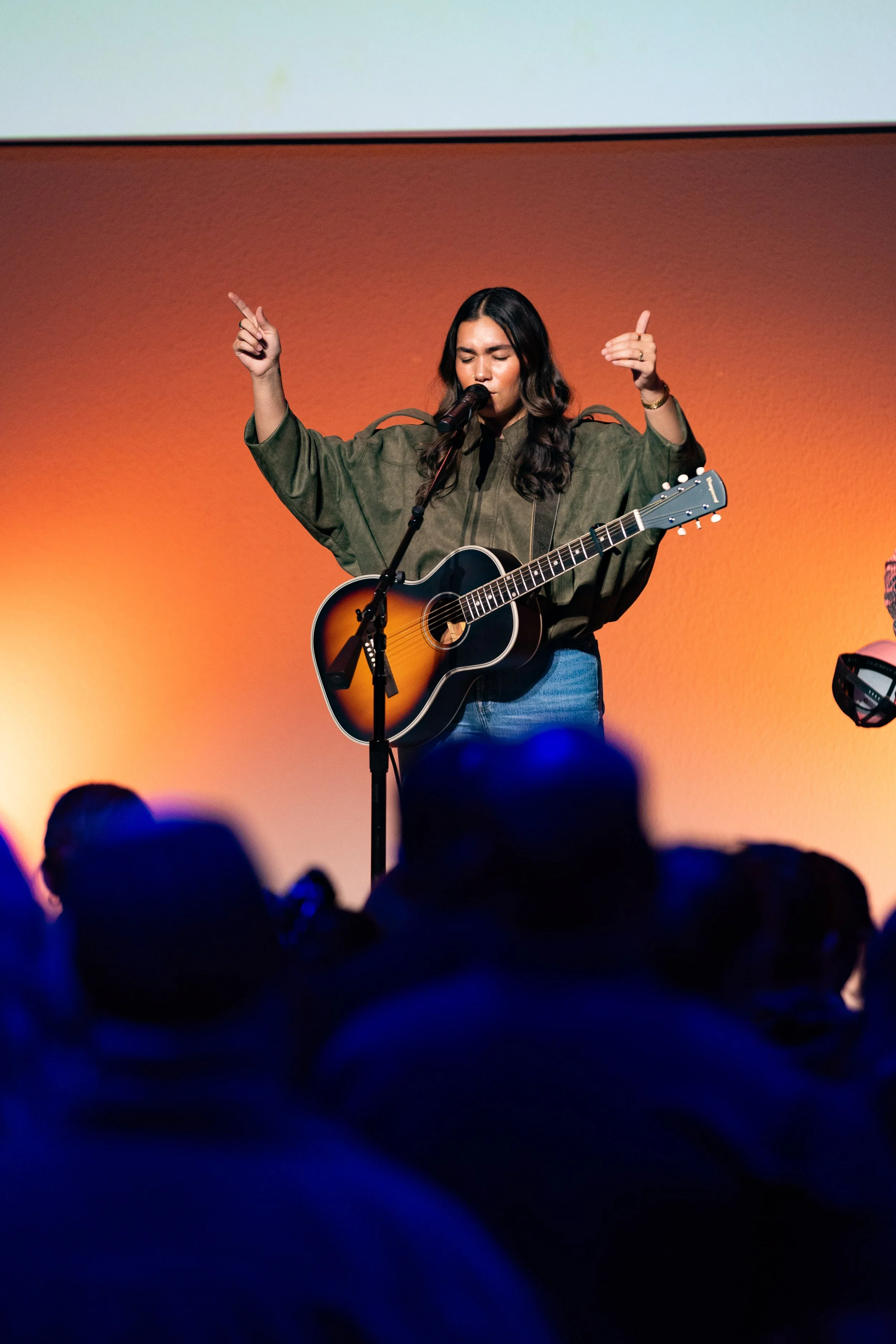 Two women singing and playing guitar on stage with a man playing guitar in the background. Large screen behind displays lyrics that say 'from the grave living in me hallelujah'.