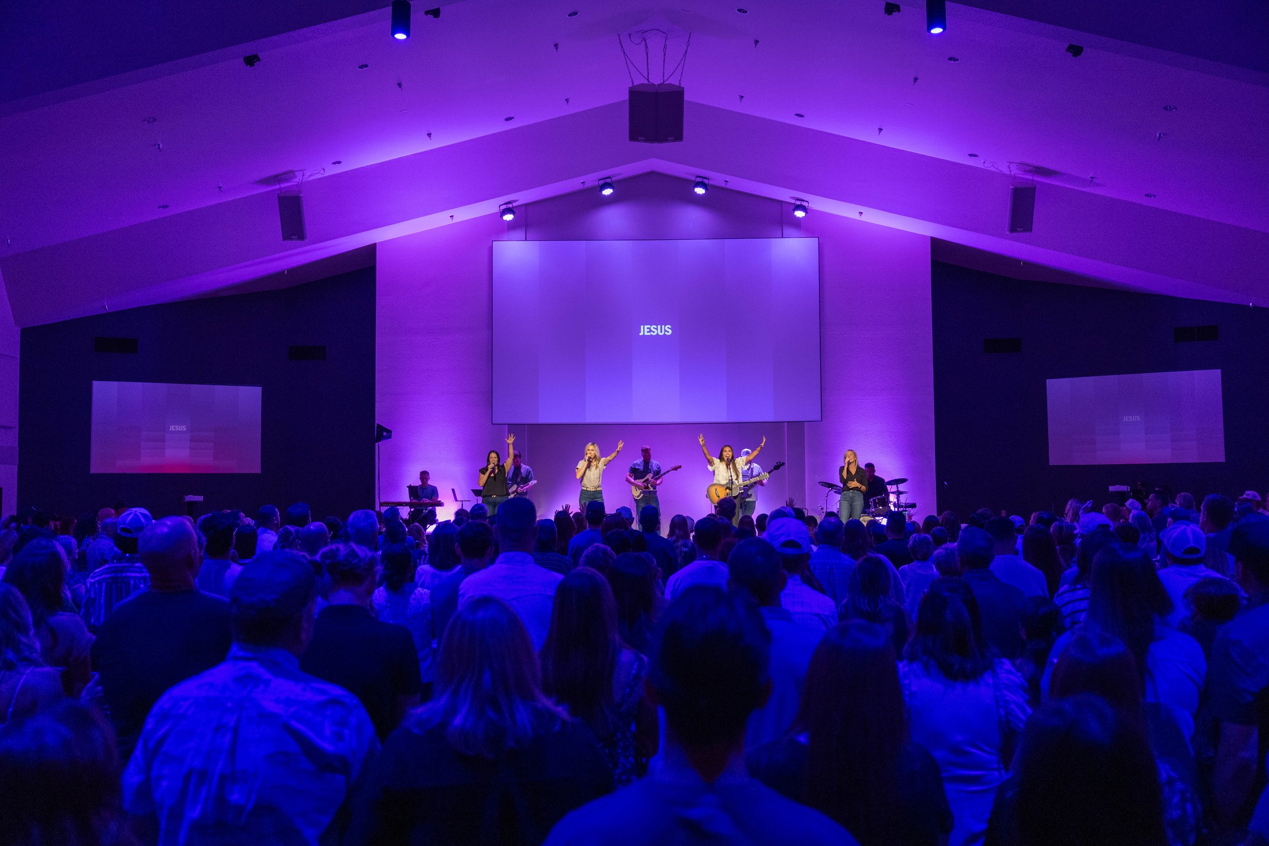 Children and adults attending a concert or performance with blue stage lighting, raising their hands or singing along as performers with guitars sing into microphones.