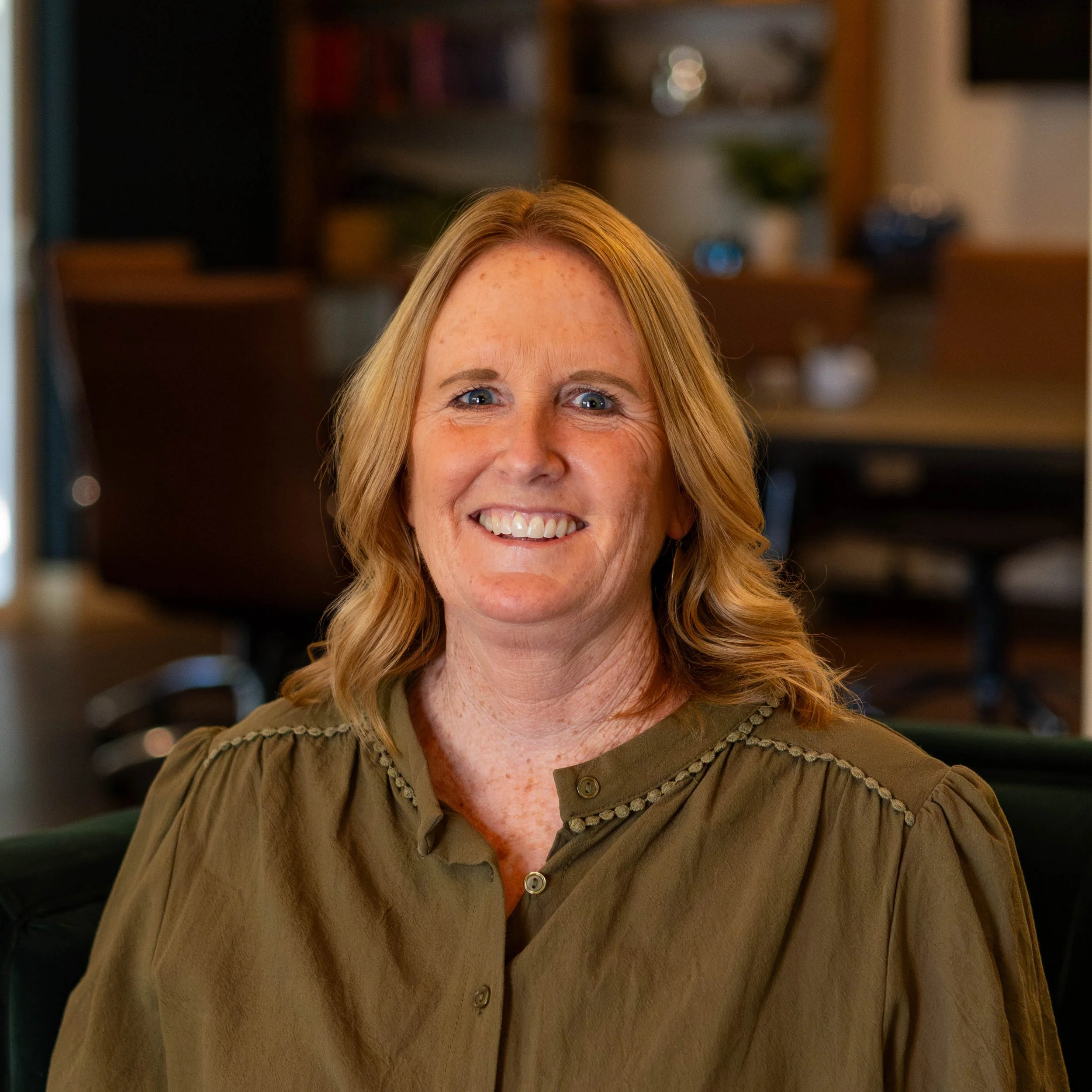 A smiling woman with shoulder-length blonde hair, wearing a black and white striped top, sitting indoors with a plant and blurred background.