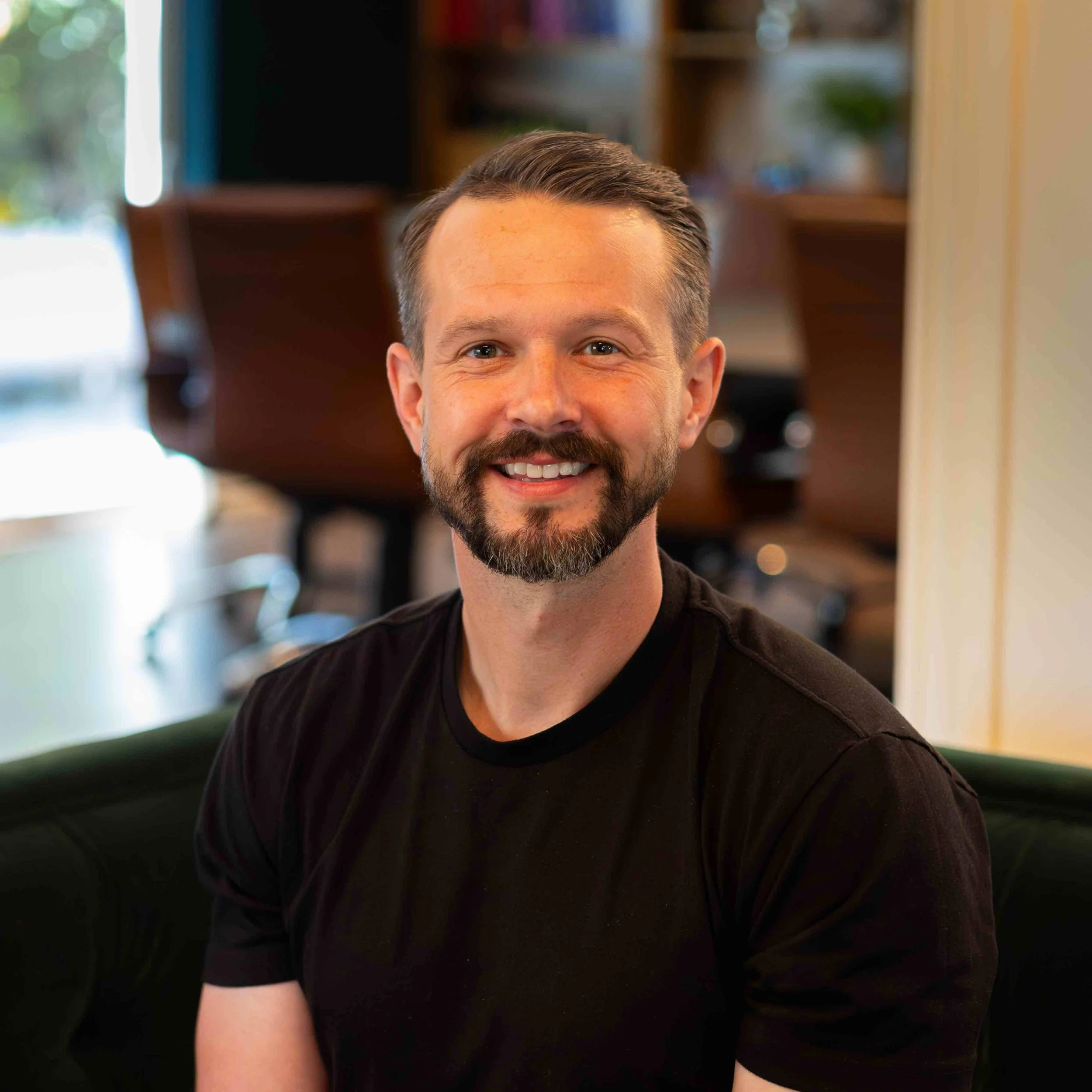 A woman with long wavy brown hair with blonde highlights, smiling, wearing a colorful patterned blouse with orange, white, and blue, and layered gold necklaces, indoors with a blurred background of plants and objects.