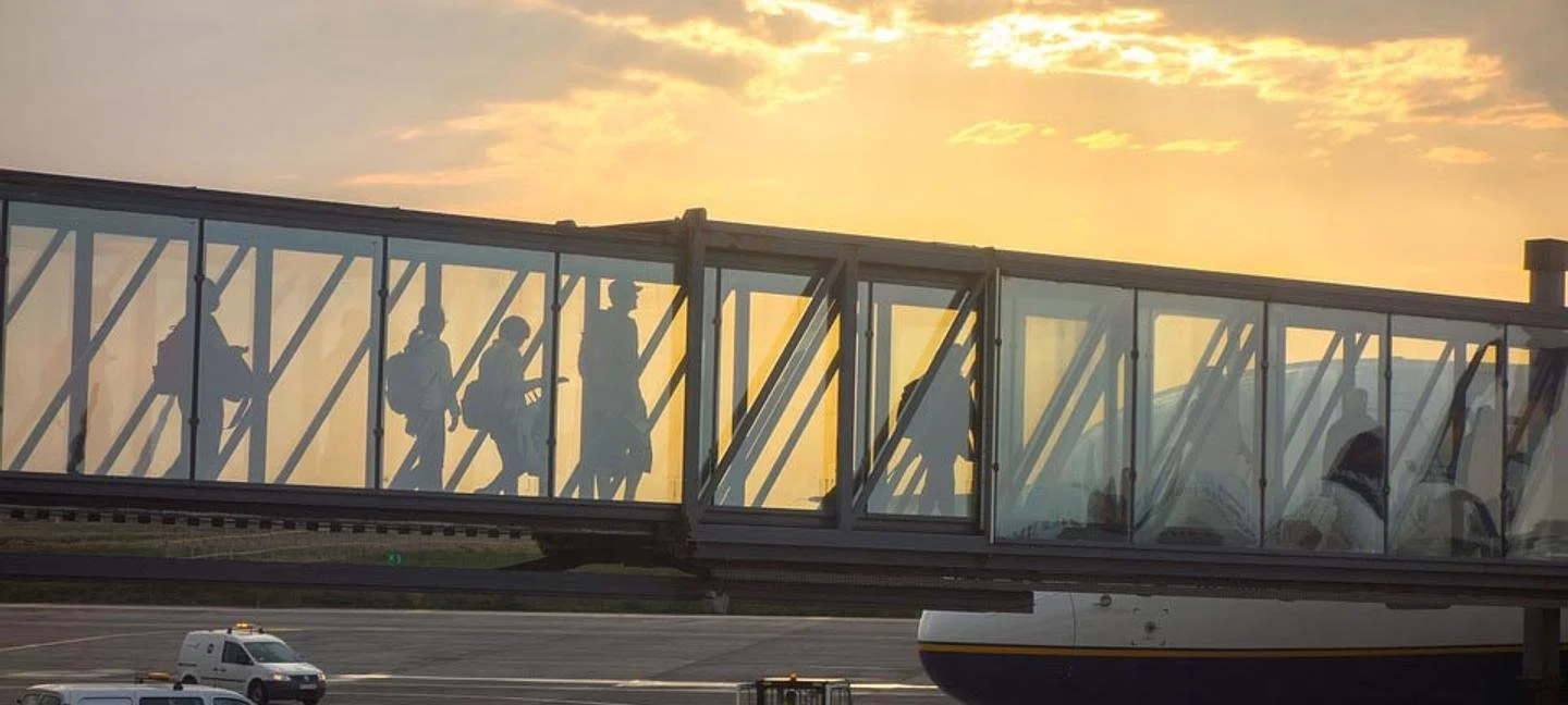 Silhouettes of travelers with backpacks walking down an airport jet bridge during sunset.