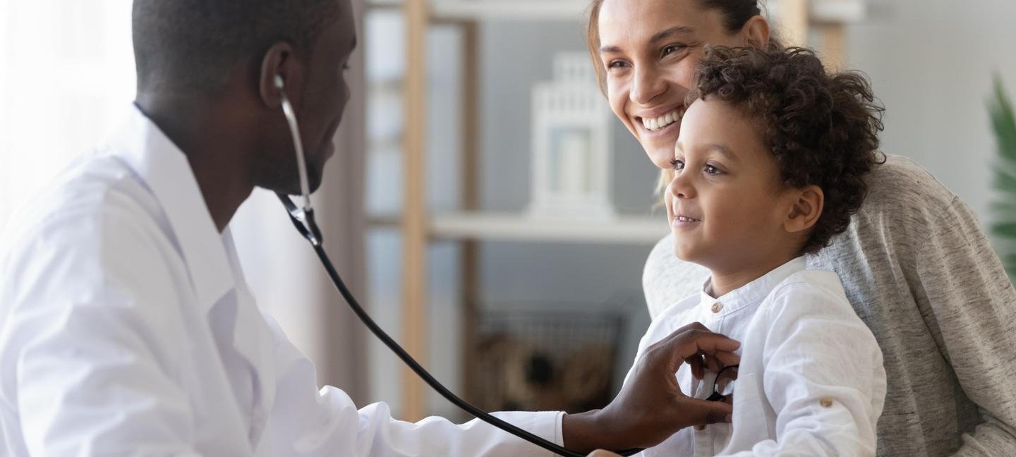 A doctor using a stethoscope to examine a smiling young boy while his mother watches happily.