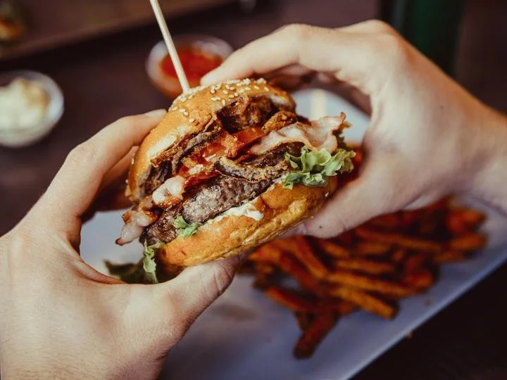 Close-up of a person holding a cheeseburger with beef patty, bacon, lettuce, tomato, and sesame seed bun, with sweet potato fries in the background.