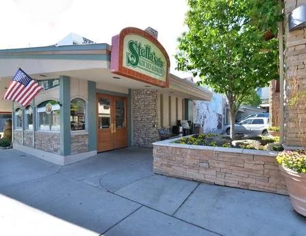 Exterior of Stella's restaurant with a stone facade, American flag, and outdoor seating area, surrounded by trees and parked cars.