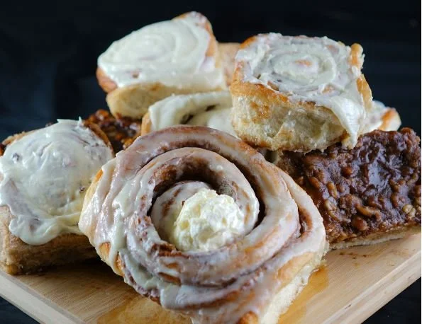 A wooden tray holds assorted cinnamon rolls, some with white icing and others with icing and filling showing.