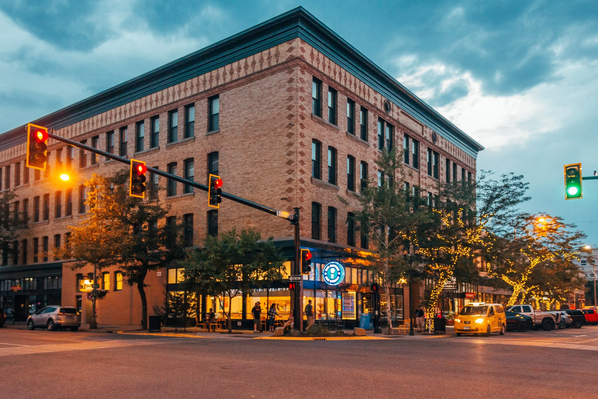 Street corner with traffic lights, a brick building, and trees decorated with string lights during dusk.