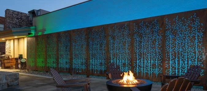Outdoor patio with a fire pit and Adirondack chairs, illuminated by colorful outdoor lighting and a decorative wooden wall with blue patterns.