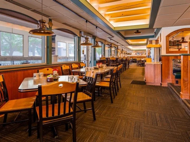 Empty dining area in a restaurant with wooden tables, black chairs, hanging pendant lights, and large windows, with a carpeted floor and a wooden partition on the right.