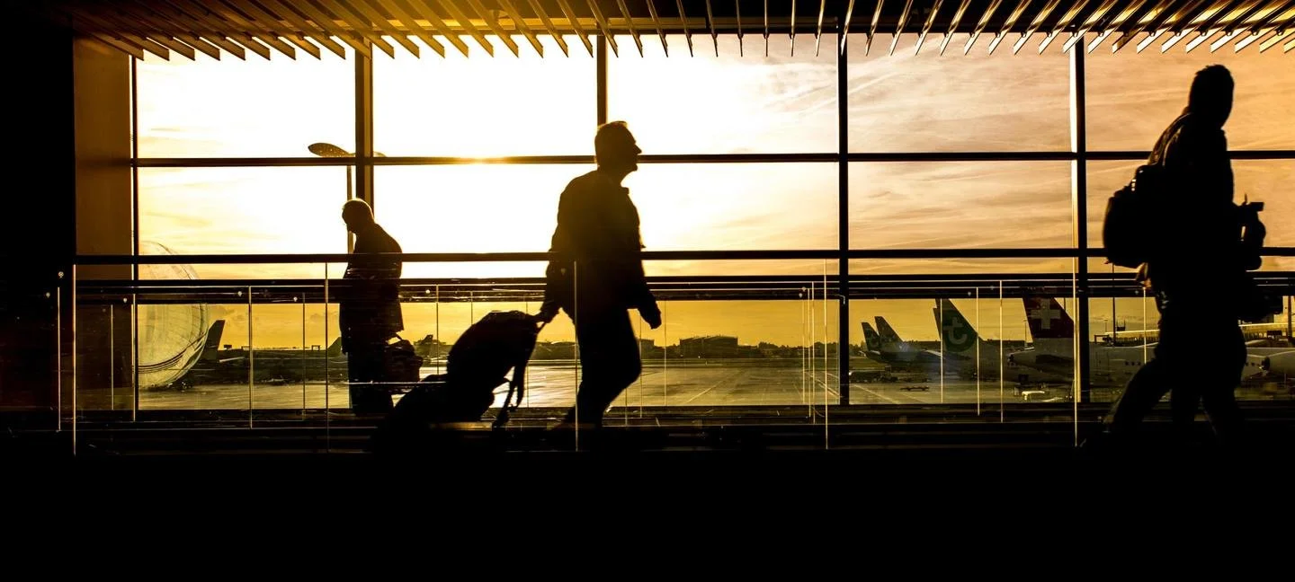 Silhouettes of three travelers walking through an airport terminal with oversized windows showing parked airplanes and a sunset sky.