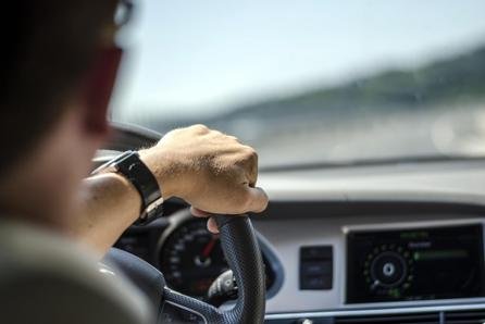 Close-up of a man driving a car, focusing on his hand gripping the steering wheel and part of the dashboard and windshield.