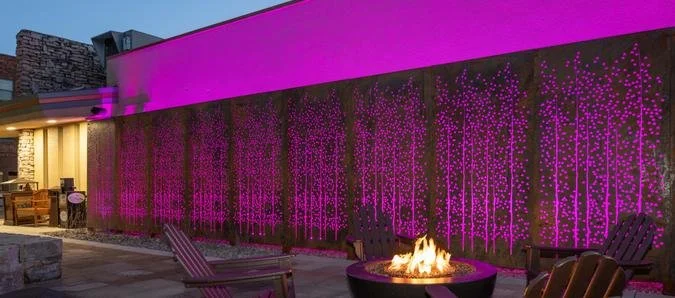 An outdoor patio with wooden chairs and a fire pit, decorated with purple string lights on a wall and purple lighting on the building