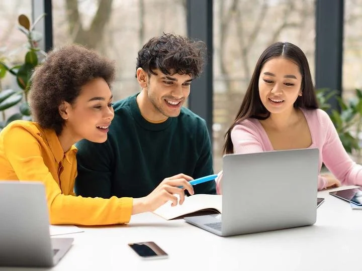 Three young adults gathered around a laptop, smiling and engaging with one another in a bright, modern setting with large windows and plants.