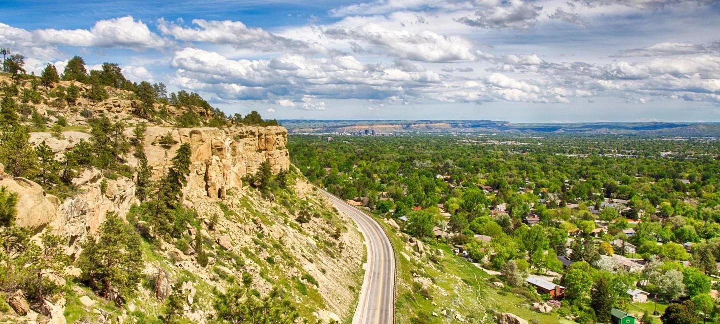 A scenic view of a winding road along a rocky, forested hillside with residential houses and a vast green landscape extending into the distance under a partly cloudy sky.
