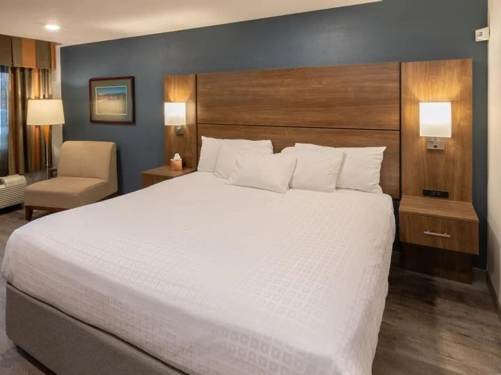 Hotel room with a large bed, wooden headboard, nightstands, wall-mounted lamps, a beige chair, a framed landscape picture, and patterned curtains.