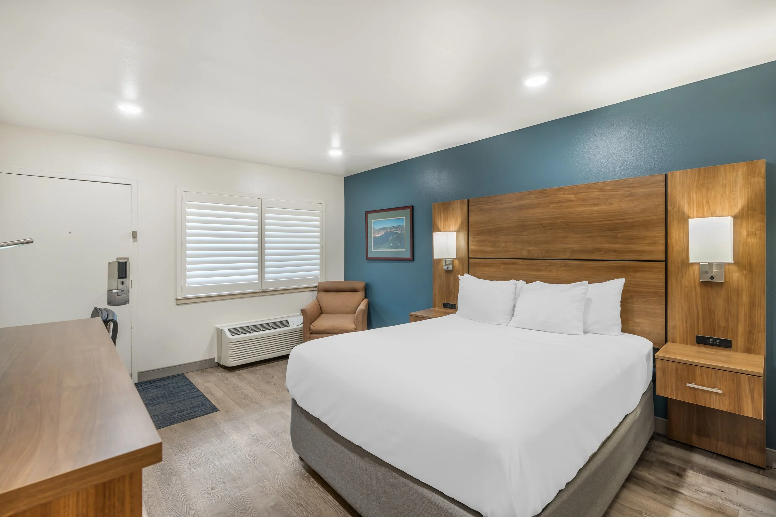 Hotel room with a large bed, wooden headboard, wall-mounted lamps, a framed picture, a beige armchair, window with white shutters, and wooden furniture.