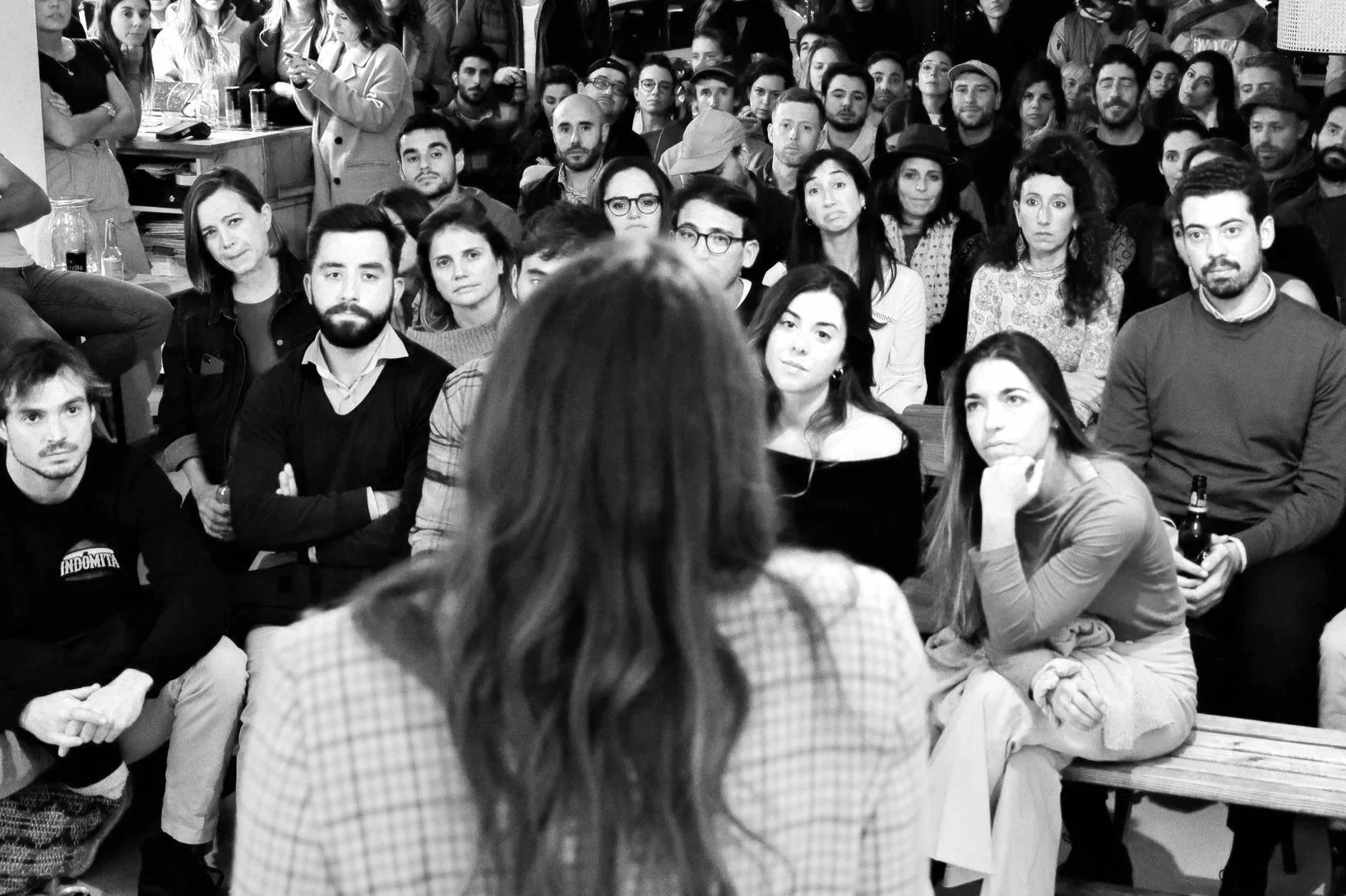 Una mujer con cabello largo y vistiendo una chaqueta de cuadros dirigiéndose a un grupo de personas que escuchan atentamente en un evento.