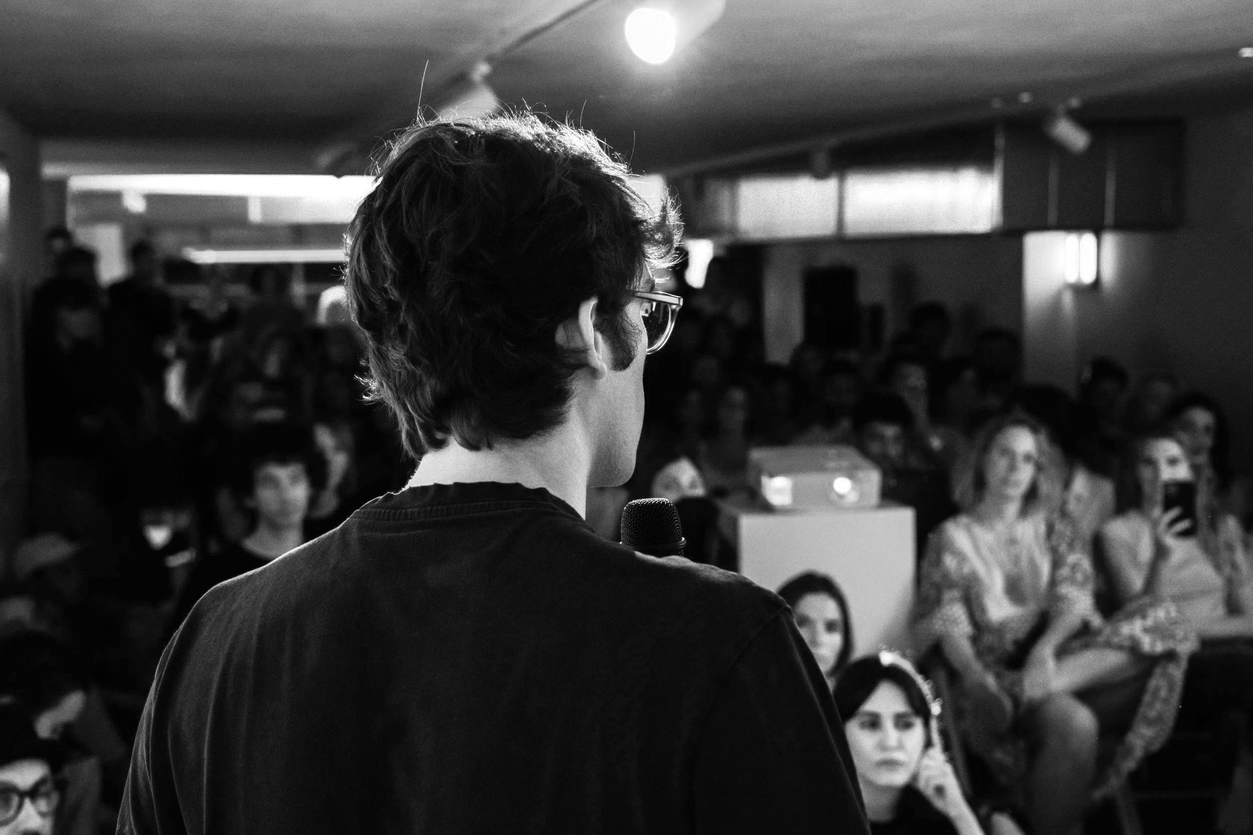 Una persona con gafas y cabello rizado sostiene un micrófono y da una conferencia o charla frente a una audiencia en un lugar cerrado.