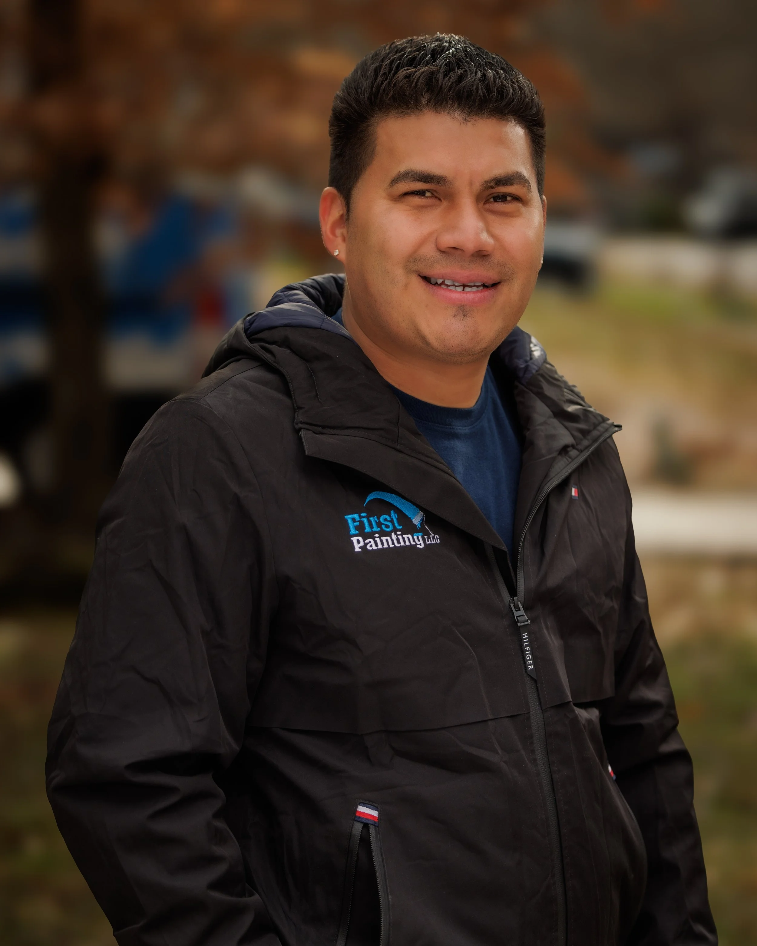 A man outdoors wearing a black jacket with a logo that says 'First Painting LLC.' He is smiling, has short dark hair, and appears to be in a park or natural setting with blurred trees in the background.