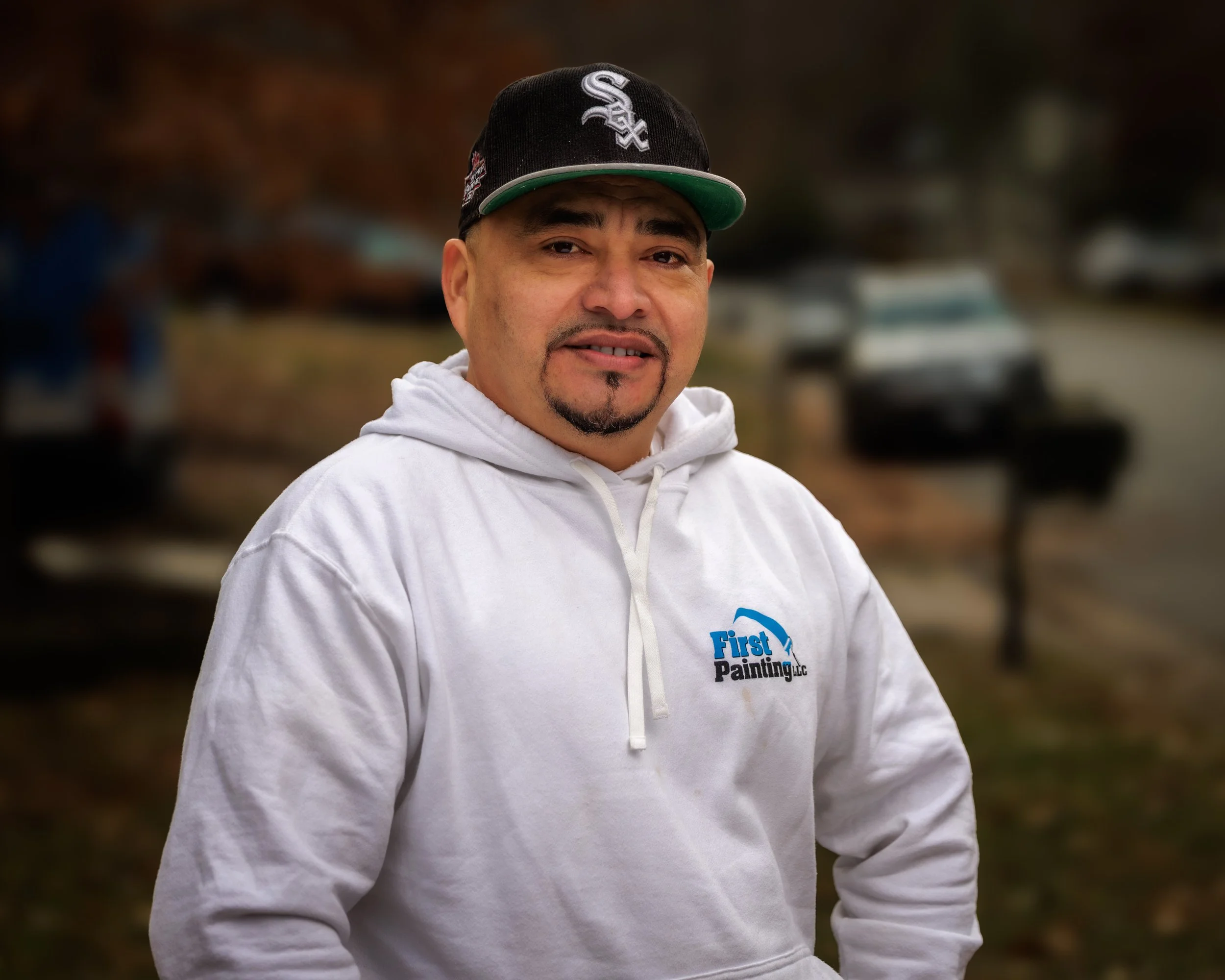 Man wearing a white hoodie with 'First Painting LLC' logo, black and green baseball cap, outdoors, with blurred cars and trees in background.