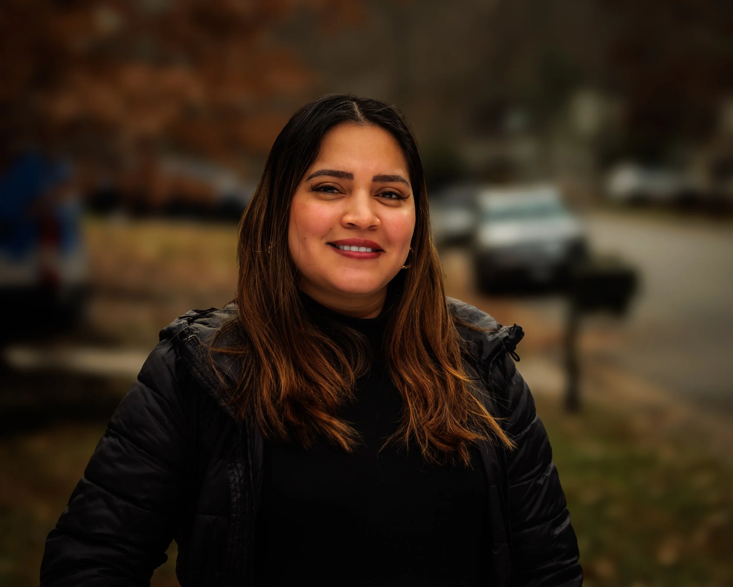 A woman with long brown hair, smiling, dressed in black, outdoor setting with blurred cars and trees in the background.
