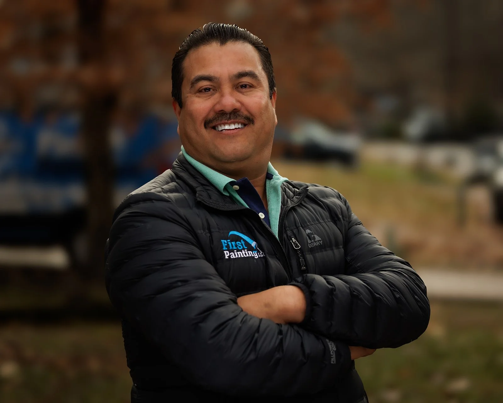A smiling man with a mustache wearing a black jacket with a logo that says "First Painting LLC" and arms crossed, standing outdoors with trees and parked cars in the background.