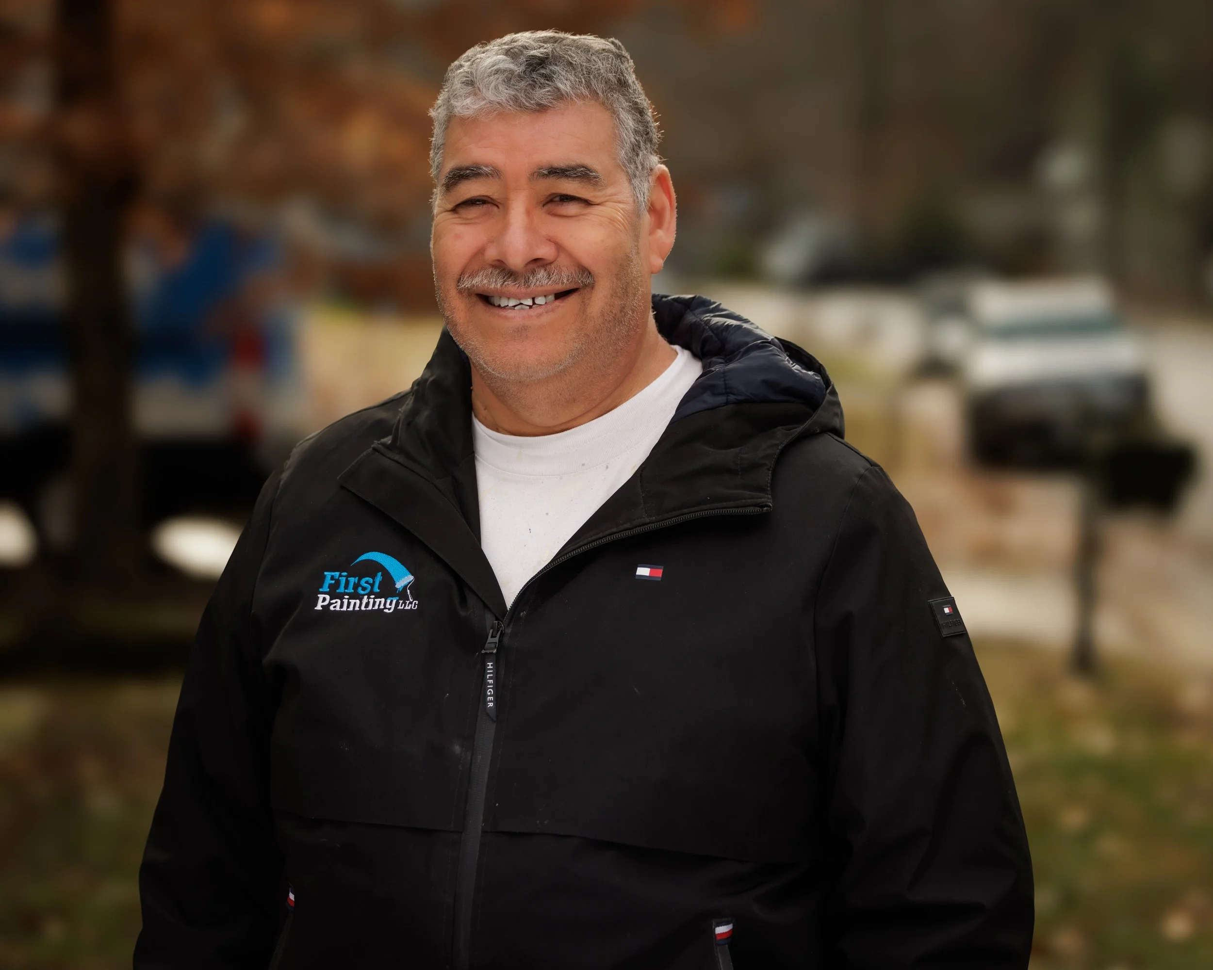 Smiling middle-aged man with gray hair and beard wearing a black jacket with 'First Painting LLC' logo, outdoors in fall.