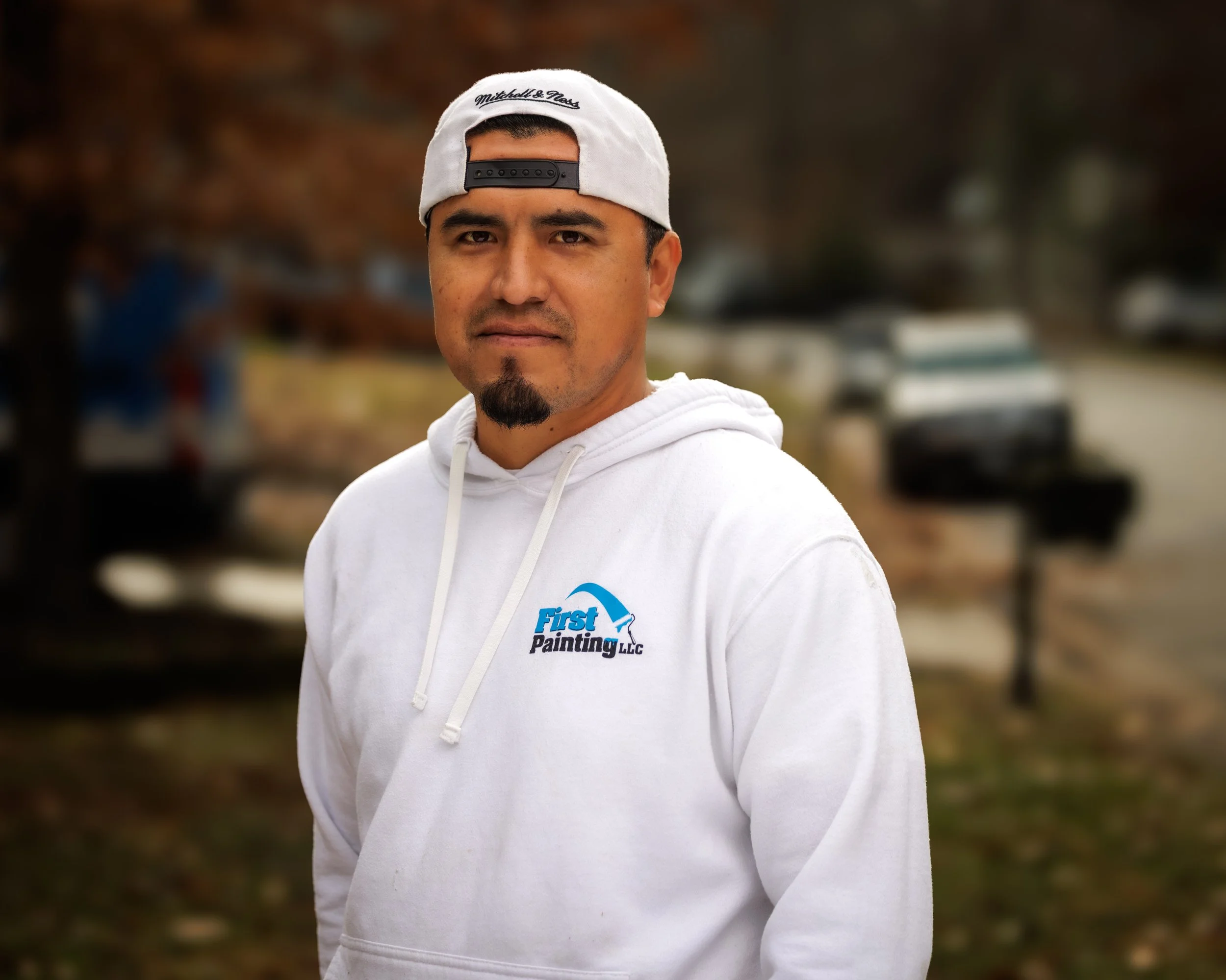 Man wearing a white hoodie and backward white cap with faded hair, standing outdoors during fall with trees and parked cars in the background.