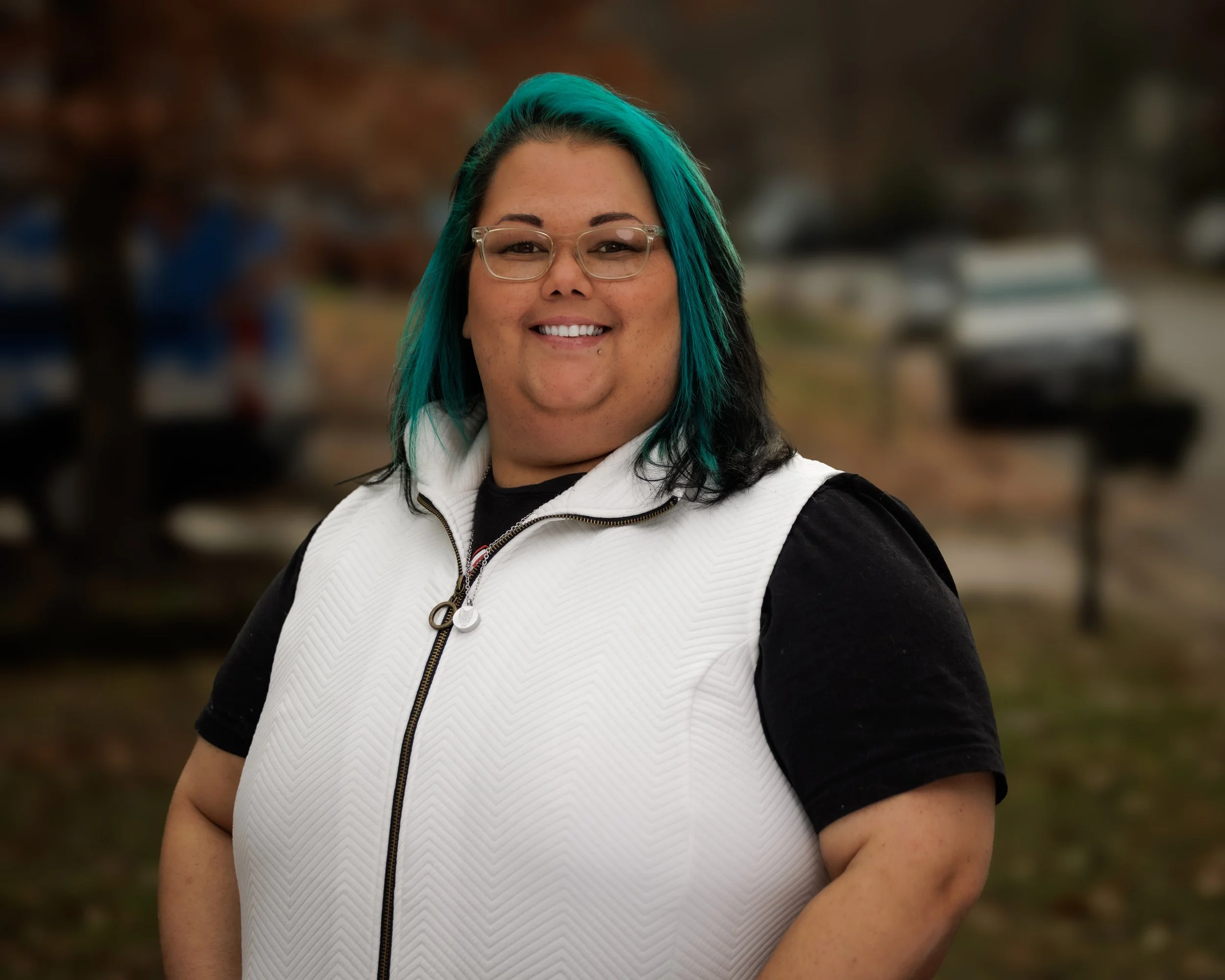 A person with teal and black hair, glasses, smiling, wearing a white vest over a black shirt, standing outdoors with trees and parked cars in the background.